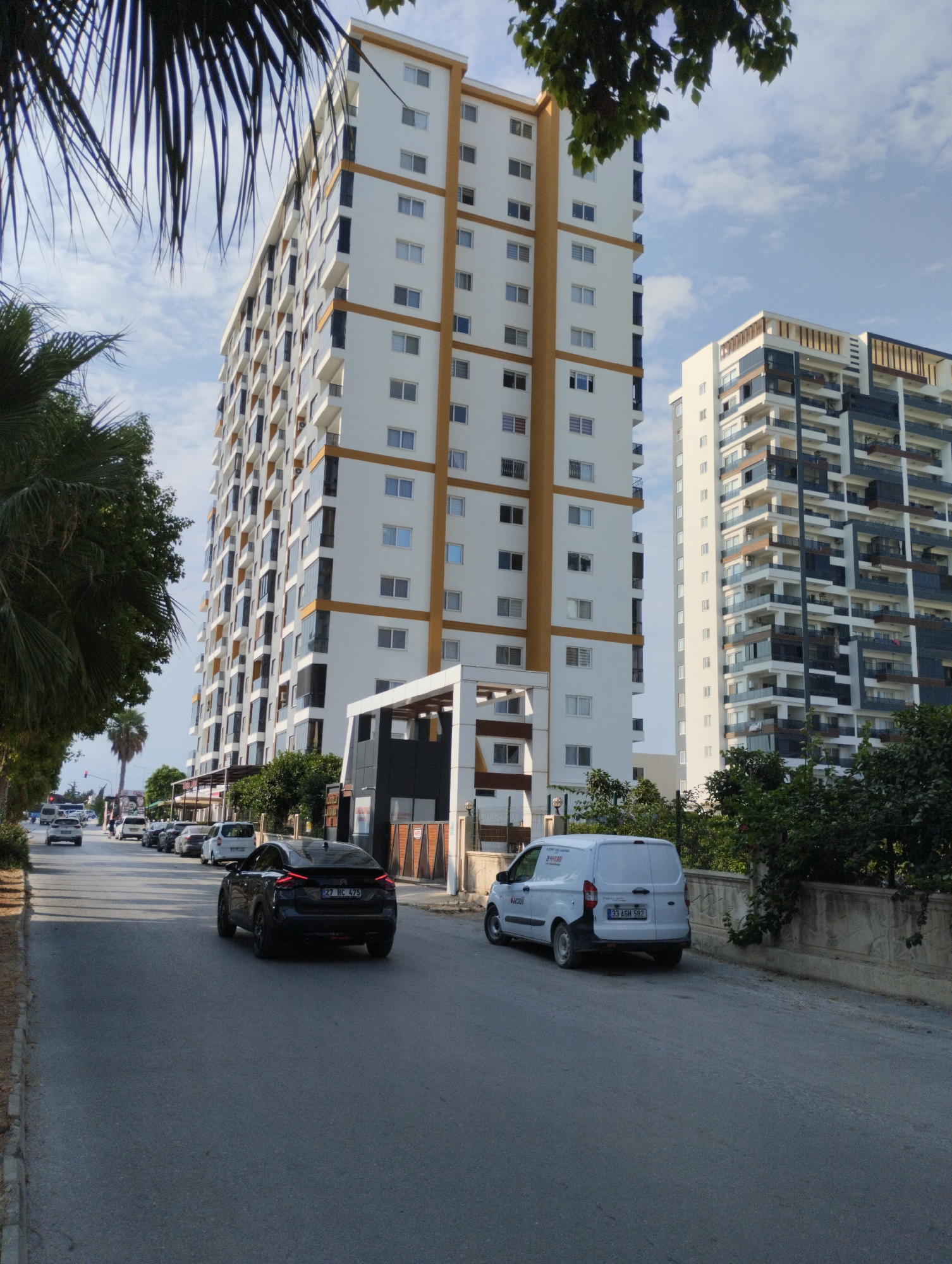 In Erdemli, Turkey, two modern high-rise apartment buildings dominate the skyline, one significantly taller than the other, with both featuring white exteriors accented by balconies and windows. The taller building has a distinct orange vertical banding, while the shorter one displays a pattern of dark blue and white. In the foreground, a paved road runs through a city environment. A dark-colored car with license plate "27 HC 475" is driving away from the viewer on the road, while a white van with license plate "33 AGH 582" is parked on the side of the road. Palm trees and leafy trees line the left side of the road, partially obscuring the view of the buildings and the sky, which is partly cloudy. In the background, several other cars are visible on the road, suggesting ongoing traffic. Shops and smaller buildings are situated along the street between the trees and the apartment complexes.