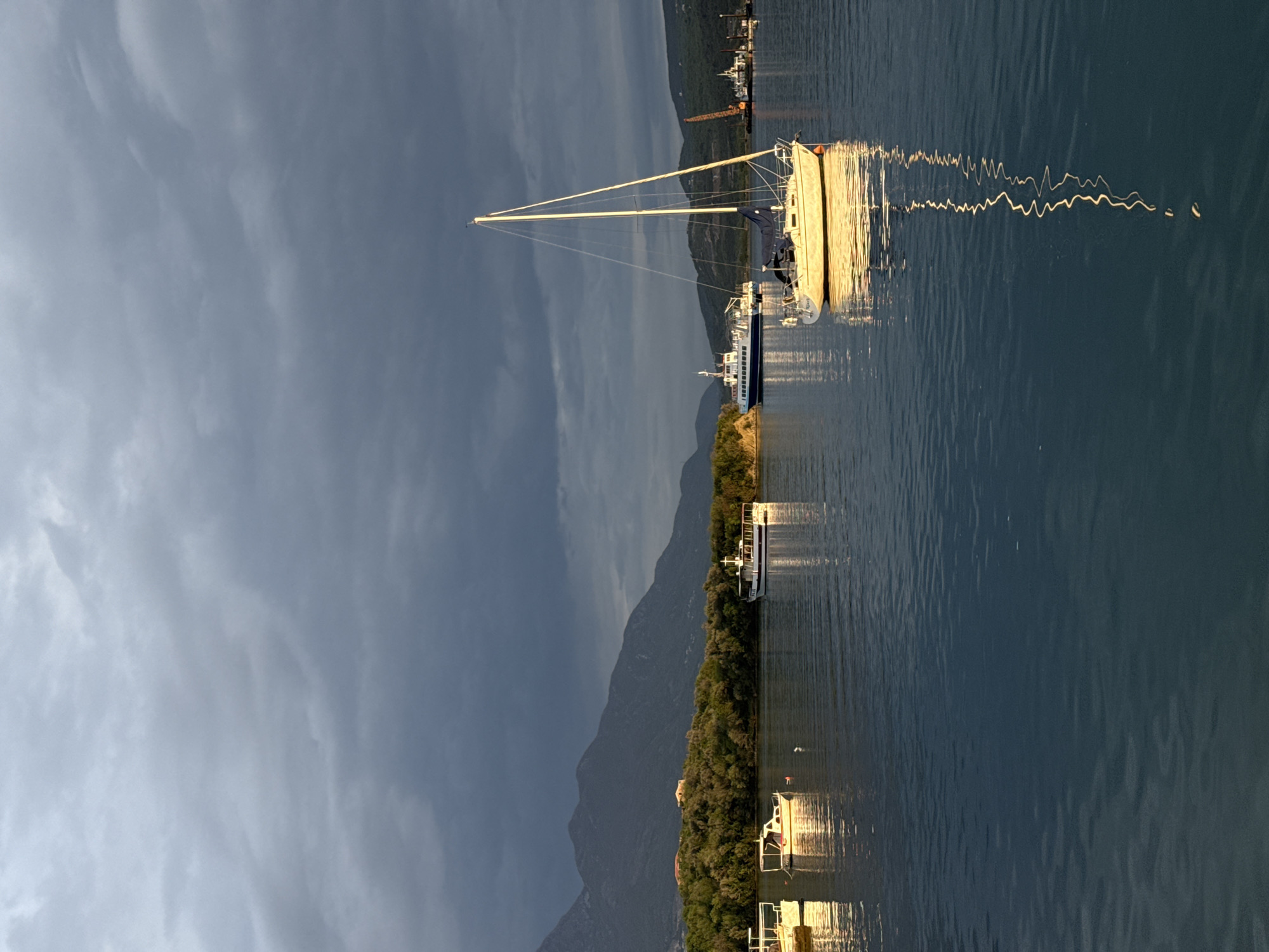 A landscape oriented photograph depicts boats in a body of water, potentially a harbor or bay, under a dramatic sky. The image is most likely taken in Bogišići, Montenegro, given the setting. The main subject is a sailboat with a tall mast, its hull reflecting sunlight, and other vessels are visible nearby. A forested shoreline runs along one side of the water, backed by distant mountains partially obscured by clouds. The sky features a mix of dark and light clouds, suggesting either an approaching storm or clearing weather. The water is calm with slight ripples that reflect light and create patterns. Several buildings are located along the shoreline. The presence of docked boats and buildings indicates that the photograph was taken in a settled coastal area, likely a marina or port.