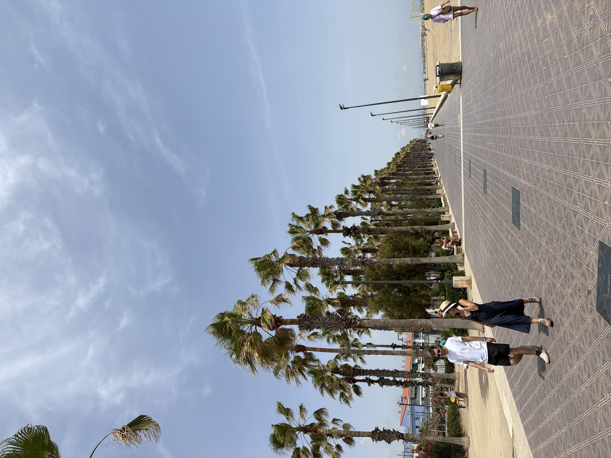 In this outdoor, daytime shot, a couple strolls along a patterned, paved walkway lined with palm trees. The man, wearing a white t-shirt and black shorts, walks on the left, while the woman in a dark blue dress and straw hat walks to his right, holding her hand to her face. The walkway stretches into the distance, leading towards a beach with a volleyball net. The sky is blue with scattered clouds, and the overall atmosphere suggests a sunny day.