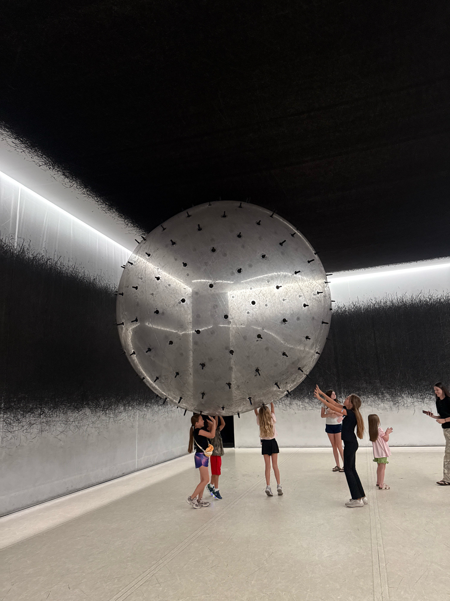 In Chicago, Illinois, USA, at the Museum of Contemporary Art Chicago, a large, metallic, spherical art installation hangs from the ceiling. The sphere is covered with numerous small, dark protrusions, resembling nails or screws, and it reflects the surrounding environment. The walls of the museum are painted black with a textured pattern of fine, dark lines, and a strip of bright white light runs along the top of one wall. Several children, appearing to be between the ages of 6 and 13, are gathered around the sphere, some of them touching or holding it up with their hands. One child on the far right, and a woman in the background on the far right, appear to be adults, possibly chaperones or museum staff. The floor is a light-colored, smooth surface with faint lines indicating seams or pathways. The overall atmosphere suggests an interactive art exhibit in a modern museum setting.