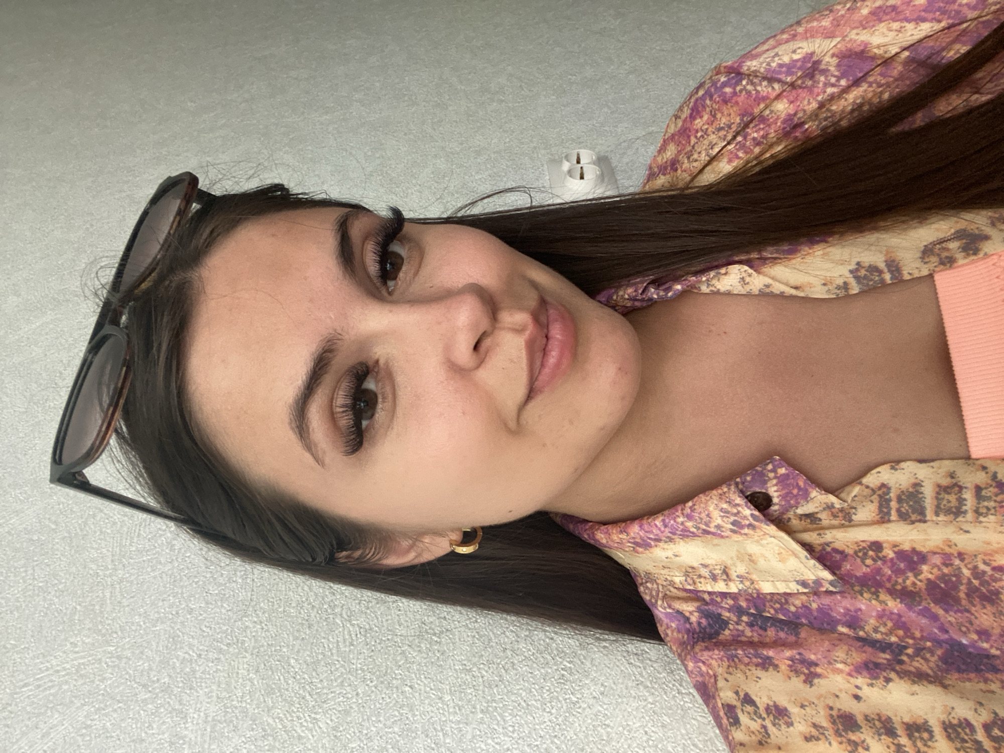 The image shows a close-up, chest-up shot of a young woman with long, dark brown hair. She is wearing sunglasses perched on her head, gold hoop earrings, and a brightly patterned button-up shirt with purple and yellow hues. She has long, voluminous eyelashes and is smiling subtly. A bright orange or pink garment is visible beneath her shirt. The background is a plain, textured grey wall with a white electrical outlet visible on the right side. There is no indication of the city of Chaikovsky, Russia, or any specific urban elements in the image.