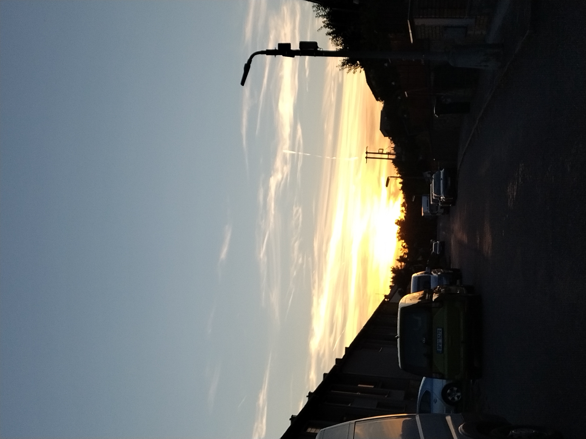 The image depicts a street scene in Dobev, Czechia, during sunset. A clear sky with wispy clouds stretches above, transitioning from light blue to a warm gradient of yellow and orange near the horizon. A prominent streetlamp with a modern design stands on the right side of the frame, silhouetted against the sky. The street is lined with parked vehicles, including a green van in the foreground with the license plate "SP9 5278" visible. To the left, a dark-colored building with multiple windows is visible, casting shadows. Trees and other structures are silhouetted against the vibrant sunset on the right side of the image. The overall impression is of a quiet, residential street at dusk.