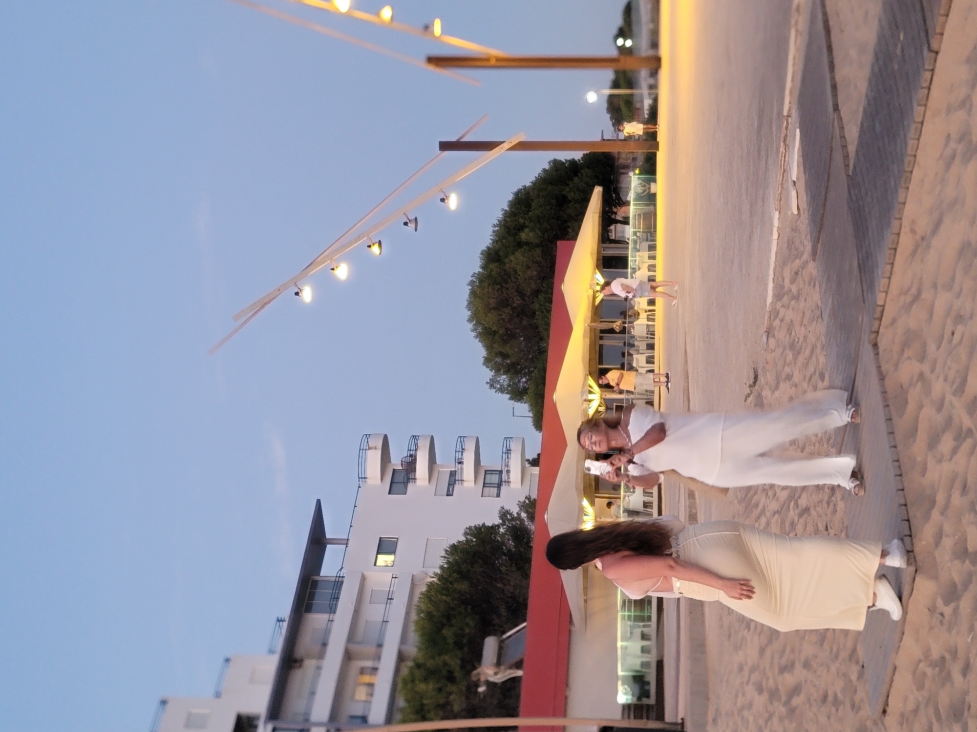 This is a vertical, eye-level, outdoor shot captured at dusk in Quarteira, Portugal. The scene features a beach with sand and wooden boardwalks in the foreground. In the midground, two women are visible; one on the left wears a light-colored, form-fitting dress and is facing away from the camera, while the woman on the right, in white pants and an off-the-shoulder top, is taking a photo with her phone. Behind them, a restaurant or cafe area with white umbrellas and tables is situated. Further back, a modern, white multi-story building with balconies is prominent. Trees are visible between the building and the restaurant. Overhead, strings of lights with yellow bulbs hang on poles, suggesting an evening setting. The sky is a clear, light blue.