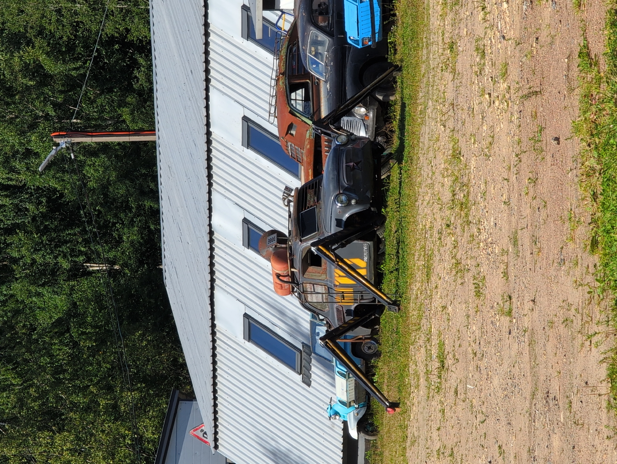 In Novoe Devyatkino, Russia, an outdoor scene captures an unusual arrangement of vehicles parked on a gravel lot in front of a long, corrugated metal building with several slanted skylights. A light blue and white scooter is parked to the left. In the center, a dark grey car has been modified with what appear to be spider-like mechanical legs, and it is elevated by these legs, with one leg on the far left partially visible. A large orange cylindrical tank is mounted on the roof of this car, along with some metal grating. To the right of this vehicle, a rusty brown car is parked, followed by a black car, and then a bright blue car. The background is dominated by dense green trees under a clear blue sky. Power lines and a wooden pole with a light fixture are also visible. The ground is covered with gravel and patches of green grass.
