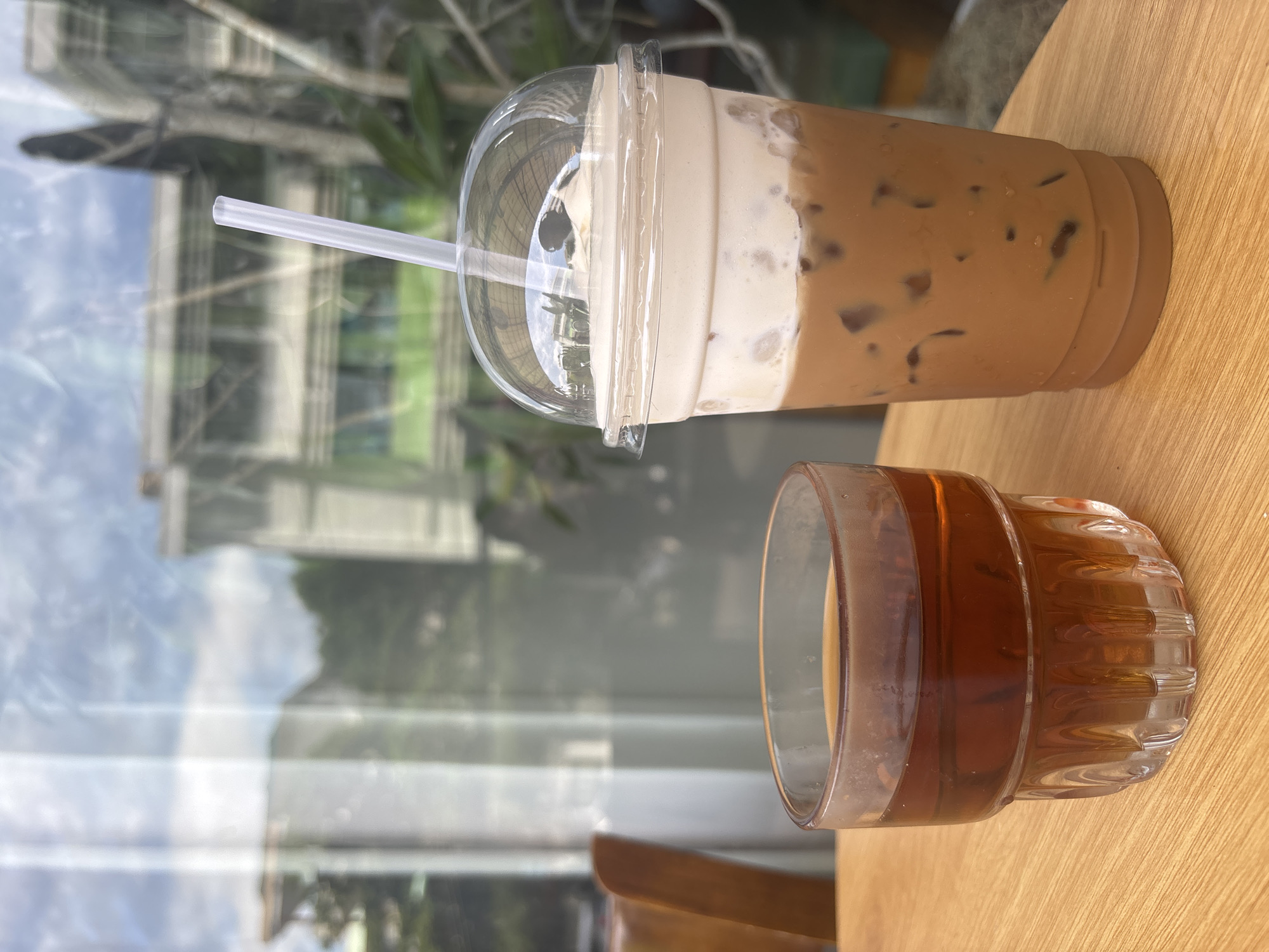 Two drinks sit on a wooden table in front of a window that reflects a building and trees. On the left is a clear glass filled with a dark amber liquid, possibly tea or iced coffee. On the right is a clear plastic cup with a domed lid and a white straw, containing a creamy, layered beverage. The lower portion of the drink is a light brown, suggesting coffee with milk, and is filled with ice cubes. Above this is a thick layer of white cream, topped with a small dollop of whipped cream. The background through the window is blurred, showing the architectural details of a building and lush green foliage. A wooden chair is partially visible on the far left of the frame.