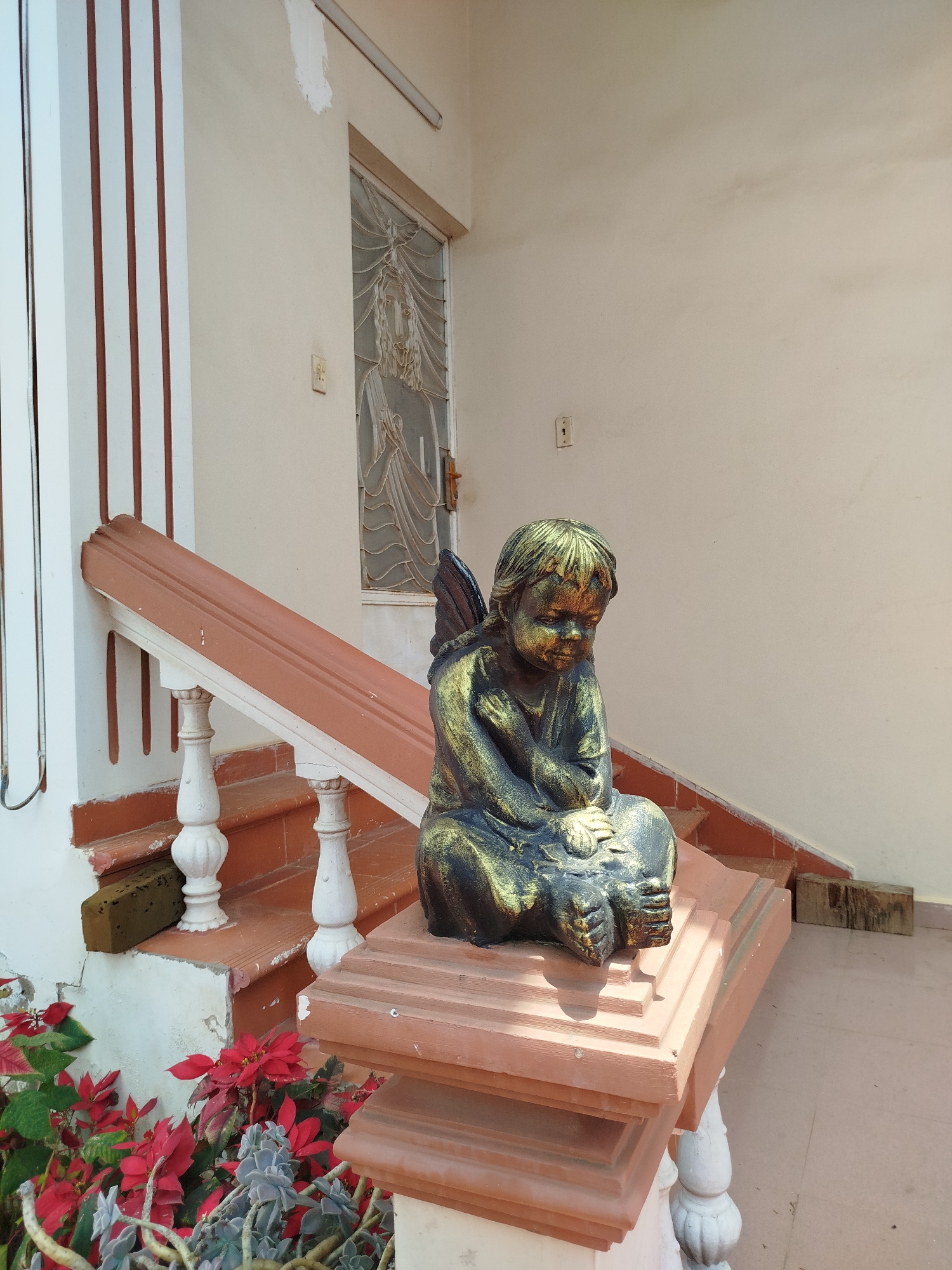 An outdoor scene captures the entrance or porch area of a building in Lâm Viên, Đà Lạt, Vietnam, under bright daylight. A statue of a young figure, resembling a pensive cherub or angel, sits prominently on the square newel post at the base of a staircase railing in the foreground. The statue is dark with distinct gold highlights on its hair, face, and clothing, and its hands are clasped in front.
The staircase features terracotta-colored treads and risers, with a matching brown handrail. The balusters and the newel post supporting the statue are painted white. The surrounding building walls are a light cream or off-white color, showing some signs of age and minor wear, including a patch of lighter paint near the top right. An electrical outlet is visible on the wall.
To the left of the stairs, vibrant red poinsettia-like plants and grey-green succulents grow, adding a touch of nature and color to the built environment. In the background, a door is visible, featuring an ornate glass panel with a stylized depiction of a robed figure, possibly religious. Above the door, a fluorescent light fixture is mounted. A few wooden blocks are seen resting on the ground next to the stairs. The overall impression is a quiet, somewhat weathered domestic or religious outdoor space.