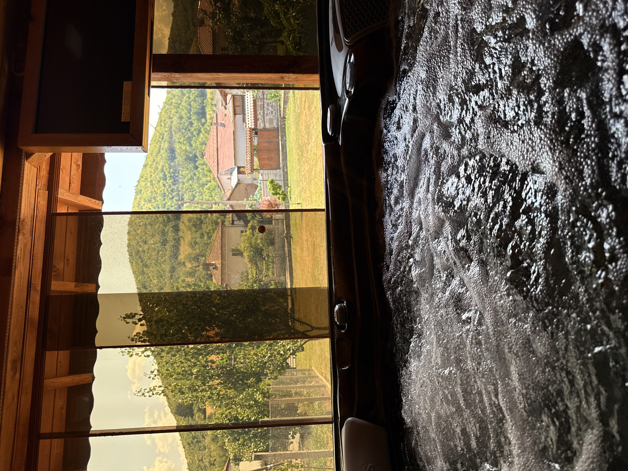 This image captures a view from inside a hot tub, looking out through a covered porch with amber-tinted windows. The foreground is dominated by the churning, bubbly surface of the hot tub water. Beyond the window, a picturesque scene unfolds in Chiprovtsi, Bulgaria. Green, tree-covered hills rise in the background, dotted with traditional Bulgarian houses featuring red-tiled roofs. A well-maintained yard with green grass and trees is visible between the houses and the porch. A television is mounted on the wooden ceiling of the porch. The overall impression is one of relaxation and scenic beauty.
