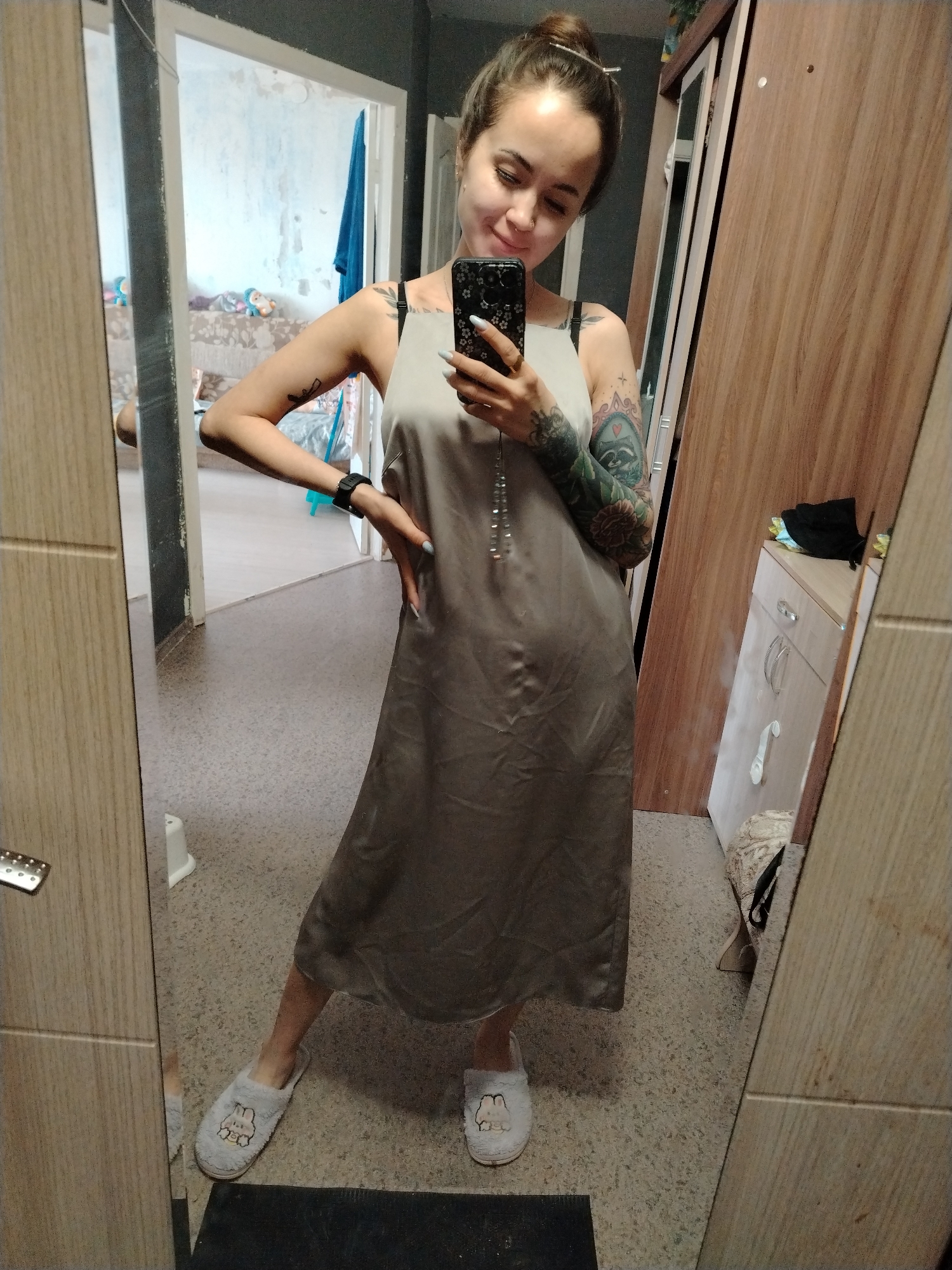 A young woman is captured in a mirror selfie within a residential interior, likely in Noviy, Russia. She stands with a soft smile, holding a black smartphone with a floral case in her left hand, angled to take the picture. Her dark hair is pulled back into a bun secured with a silver clip, and she has a nose piercing. Both her arms are adorned with extensive, colorful tattoos, including floral patterns and a depiction of a raccoon-like animal. She is dressed in a light beige/greyish satin slip dress over a dark bra, a dark wristwatch on her left wrist, and light-colored fluffy slippers featuring a cartoon bunny design.
The setting features light grey/brown speckled flooring. To the woman's right, a wooden wardrobe with a mirror reflects a light brown dresser and a black bag or item of clothing. To her left, an open doorway reveals another room with light-colored walls that appear to be partially stripped or damaged, suggesting the space might be under renovation or show signs of wear. A patterned sofa, a blue towel or fabric, and what look like toys are visible in the background room. The overall atmosphere is casual and relaxed, indicative of a moment at home. There is no clearly legible text visible in the image.