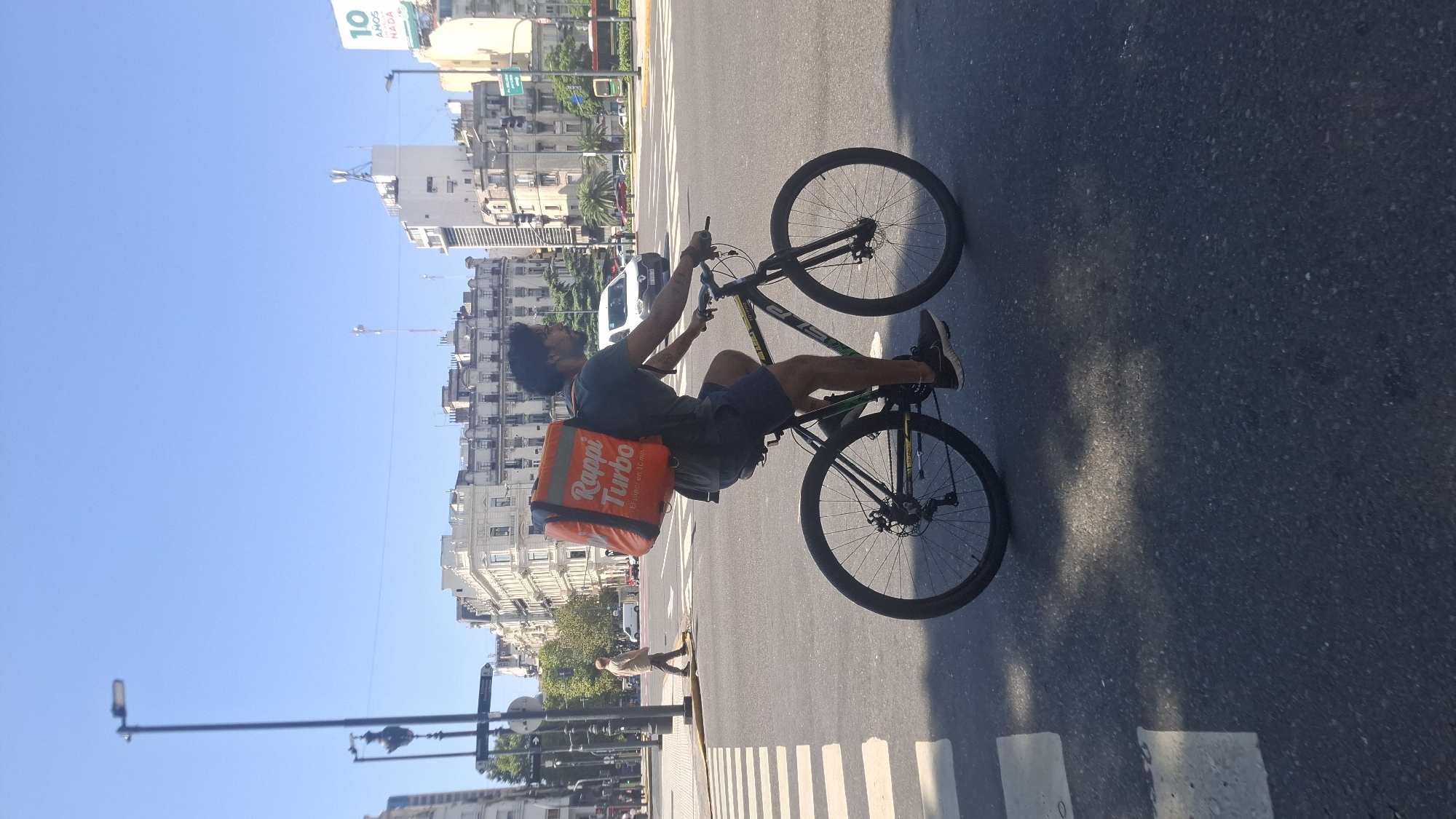 A medium shot depicts a person on a bicycle, executing a wheelie in Buenos Aires, Argentina. The individual has dark hair and wears a green t-shirt, dark shorts, and dark sneakers. An orange "Rappi Turbo" delivery bag is on their back. The bicycle is a mountain bike with black rims and a dark frame. The person is on a crosswalk on an asphalt street. Buildings and trees line the background. The sky is clear and blue.