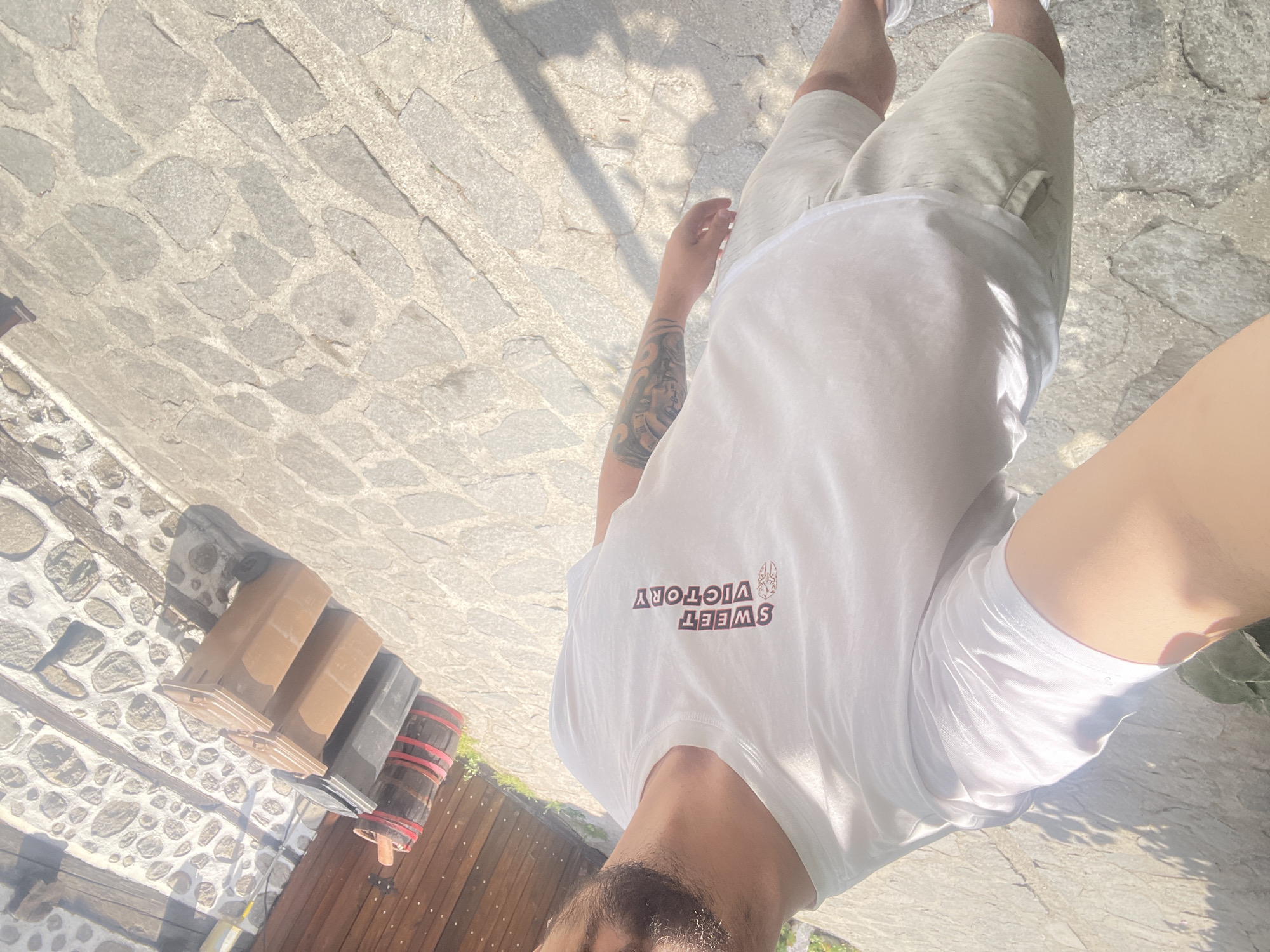 A person is taking a selfie outdoors. The person is wearing a white t-shirt with "SWEET VICTORY" printed on it, and light-colored shorts. They have a tattoo on their left arm. In the background, there are stacked trash bins, wooden barrels, a wooden fence, and a stone-paved ground. The lighting suggests it is daytime.