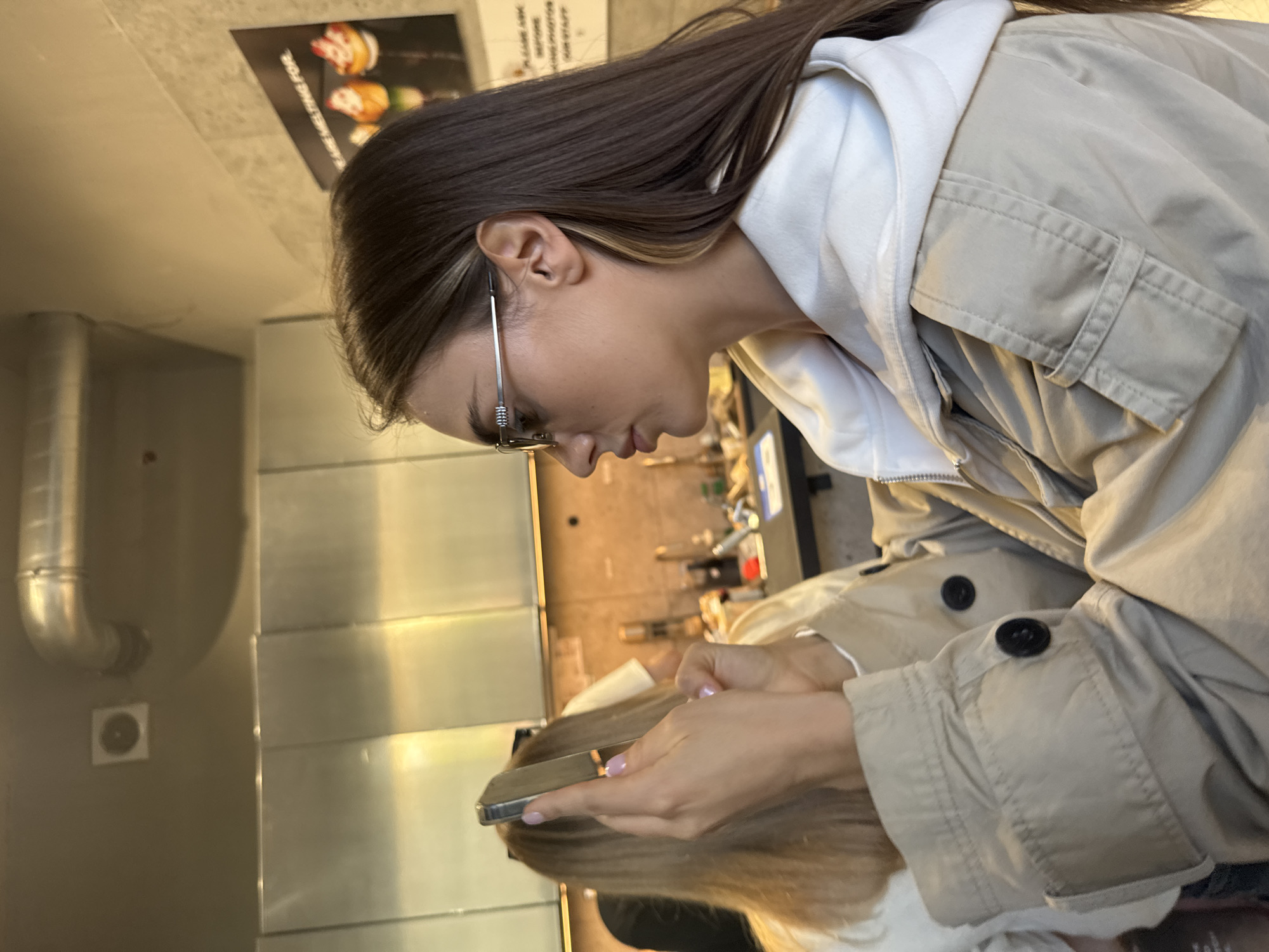 A young woman is depicted in a medium close-up, looking down at a smartphone held in both hands. She has long, straight dark brown hair with a lighter highlight near her ear and wears thin, metallic-framed glasses. Her attire includes a light beige, possibly utility-style, jacket or coat worn over a white hooded sweatshirt. Her fingernails are painted with a soft pink or lavender polish.
The woman appears to be seated or leaning over a highly reflective surface, which clearly mirrors her hair, hands, and the smartphone. In the background, there are hints of a reflection of another person or activity. The setting is an indoor space, likely a casual dining establishment such as a cafe or fast-food restaurant, suggested by the industrial design elements. To the left, a metallic exhaust pipe or vent is mounted on a plain wall, alongside a small, circular control panel. Above, on what appears to be the ceiling or an upper wall section, a dark poster displays images of food items, possibly burgers or sandwiches, with partially visible text that includes the phrase "ME TIME FOR". The lighting is warm and indirect, indicating an indoor environment. The scene captures a candid moment of someone engaged with their phone in a public, contemporary setting in Warsaw, Poland.