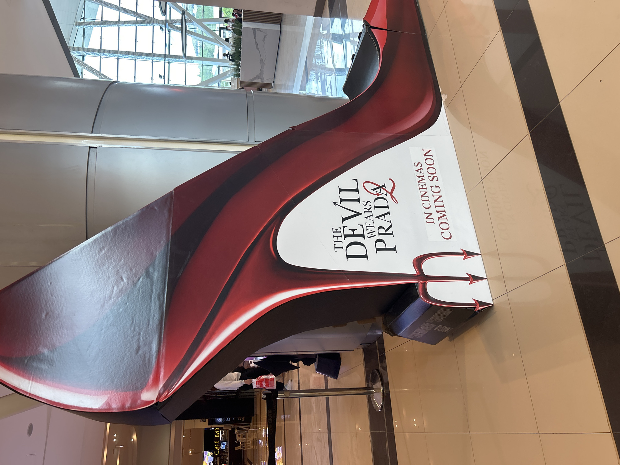 The image shows a large promotional display for the movie "The Devil Wears Prada 2: In Cinemas Coming Soon" in a shopping mall. The display is shaped like a red stiletto heel with the movie title and release information on the side. The mall has tiled floors and a modern architectural design with large windows. The exact location in Sofia, Bulgaria cannot be determined from this image alone without further contextual clues within the visible environment.