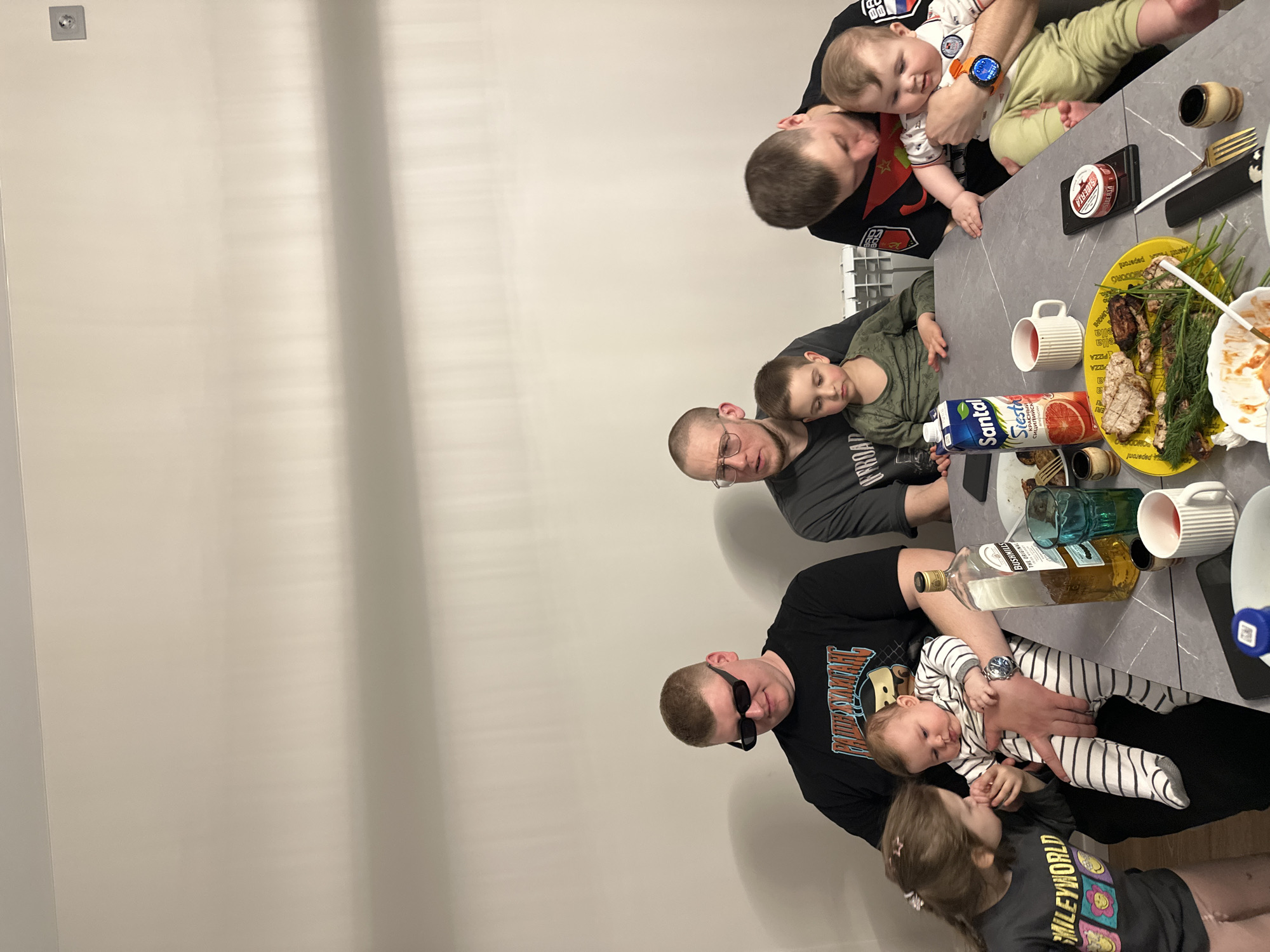 This media file depicts an indoor gathering of several individuals around a dining table, likely a family or social event in Chaikovsky, Russia. The scene includes six adult men of various ages and three children. One man, wearing a black t-shirt with "OFFROAD" visible text, holds an infant, while another man holds a second infant. A young girl, wearing a "SMILEYWORLD" t-shirt, stands next to the table.
The gray, speckled table is set with food and beverages. A prominent yellow platter holds a generous serving of grilled or roasted meat, heavily garnished with fresh dill or herbs. Other food items include a plate with remnants and a small red container labeled "Peperoni PIZZA" and "ella FUN". Drink selections include a carton of "Santal Siesta" grapefruit juice, identifiable by the Cyrillic text "СИКРАСНЫЙ СИЦИЛИЙСК" (Red Sicilian), and a bottle of "BUSHMILLS THE ORIGINAL" Irish whiskey. Several white mugs, glasses, small decorative wooden cups, and gold-colored cutlery are also on the table.
The adults are dressed casually, with another man's black t-shirt featuring "SIBERIA" text. The children appear calm and content, suggesting a relaxed atmosphere. The lighting indicates an indoor setting, possibly an evening meal. The background consists of a plain light-colored wall with visible reflections and a small wall-mounted light fixture.