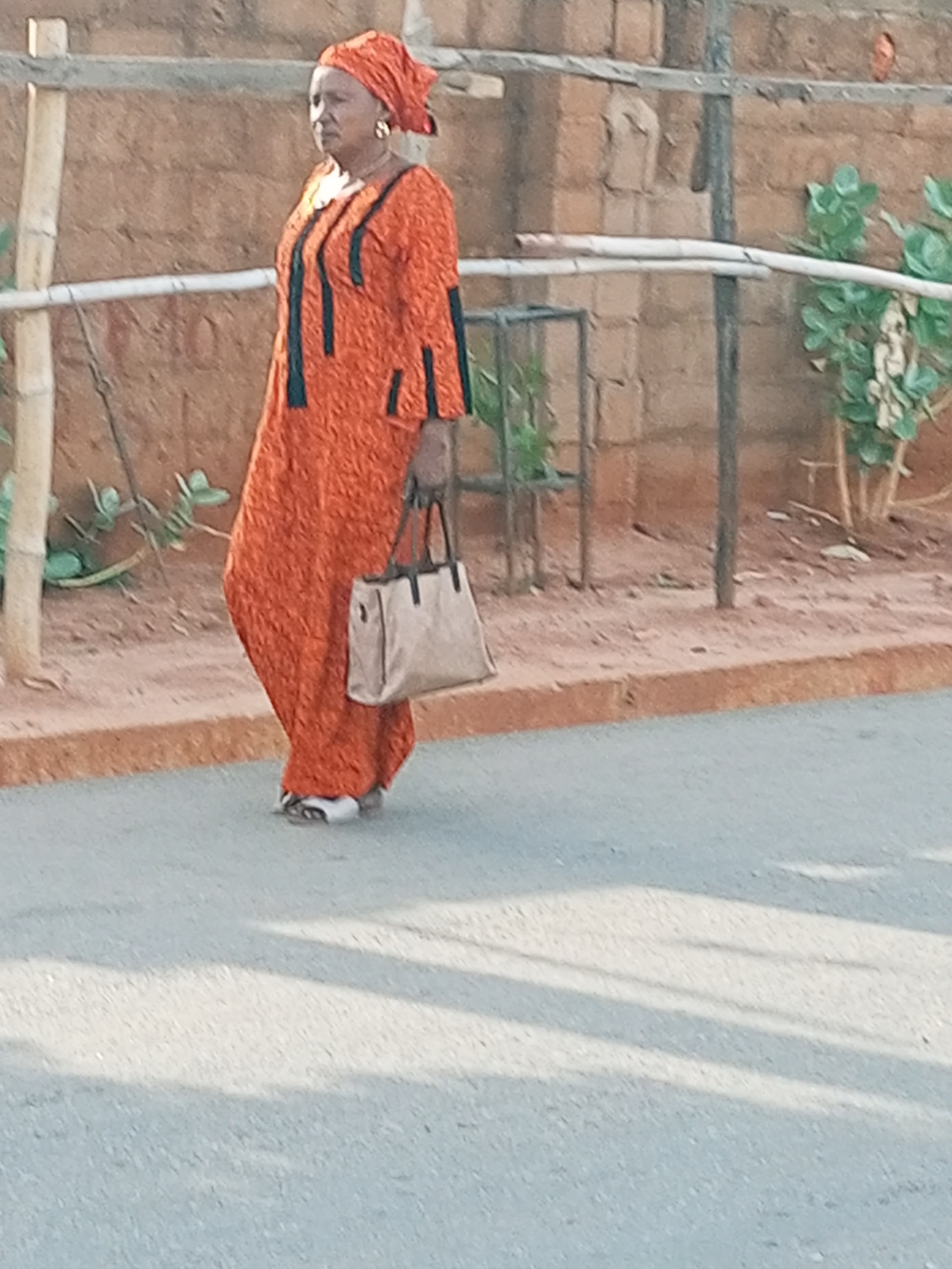A middle-aged woman, facing left, walks on a light grey asphalt road in the city of Unknown, Unknown. She wears an orange patterned headscarf, large gold hoop earrings, and a long, textured orange dress with vertical black trim on the bodice and sleeves. In her right hand, she carries a light beige handbag with dark brown straps. Her left foot is slightly raised, indicating movement. Behind her, a reddish-brown dirt shoulder borders a low, reddish-brown concrete curb. The background features a textured reddish-brown brick or mud wall, partially obscured by thin wooden or metal posts and sparse green foliage. Shadows are visible on the asphalt in the foreground.