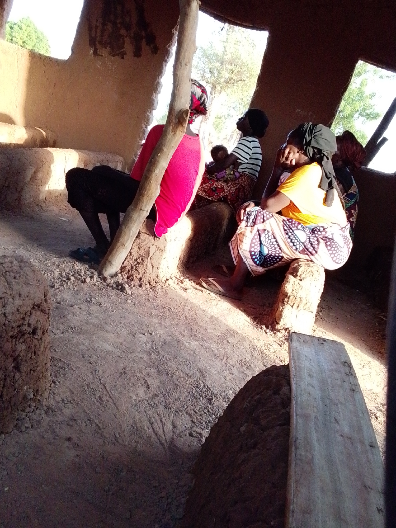 The image depicts the interior of an earth-toned structure in Mulke Barikin Sayejo, Nigeria. Four women are present. One woman, wearing a pink top and black bottoms, sits on an earthen step with her back to the viewer, leaning against a wooden support beam. Adjacent to her, another woman in a striped shirt and patterned skirt holds a baby, both looking towards the open structure. To the right, a third woman wearing a yellow top and patterned skirt sits on an earthen seat, her head covered with a dark scarf. Another woman with dark clothing and a patterned top is sitting behind the woman in the yellow top. The floor of the structure is earthen. A wooden plank is resting near the bottom right of the frame. Light streams in through the openings of the structure, illuminating portions of the scene. The overall setting suggests a rural or traditional context.