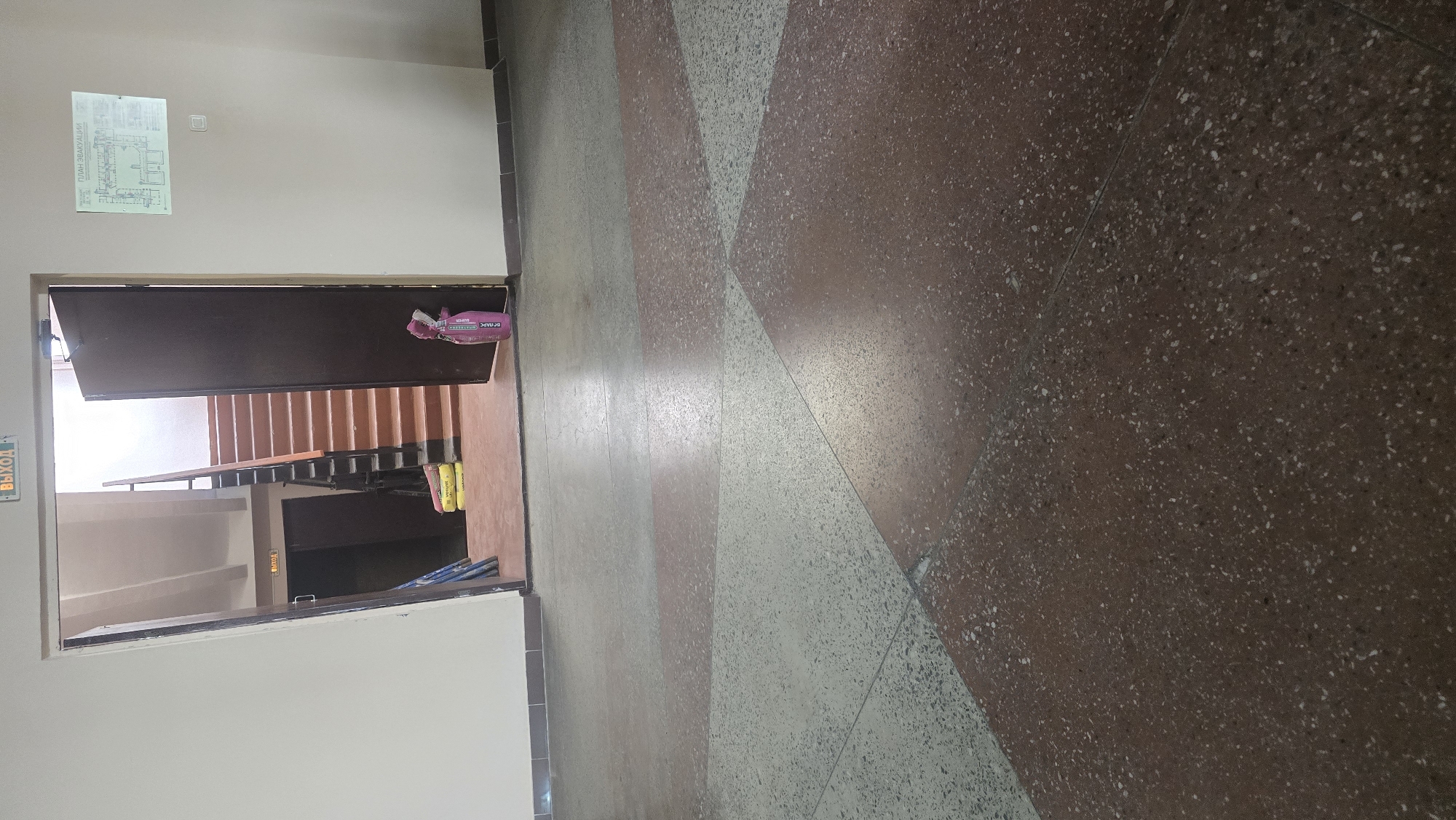 The image depicts a hallway with a staircase visible through an open door. The hallway floor is a terrazzo with a pattern of red and light gray diamond shapes. To the left, a door is open, revealing a staircase with light brown steps and dark railings. A pink duffel bag rests on the floor near the open door, and two yellow bags of what appear to be building materials are placed near the bottom of the stairs.
On the wall to the left of the doorway, a white sign with a diagram is mounted. There is also a green sign with text that appears to say "ВЫХОД" (EXIT) in Russian, mounted on the wall to the left of the doorway. Another similar green sign is visible on the wall inside the hallway leading to another door.
The lighting suggests it is daytime, possibly indoors with artificial lighting. The scene appears to be a public building, such as an apartment complex or an office building, given the presence of a staircase and exit signs. There are no people visible in the image. The overall impression is a utilitarian and somewhat dated interior space.