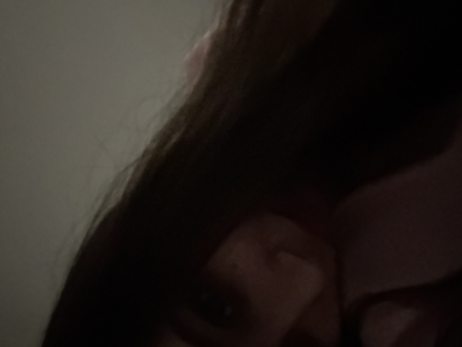A dimly lit, close-up shot focuses on a person's face, partially obscured by dark hair. One eye is visible, and a hint of nose and mouth are suggested. The background consists of a plain, lighter-colored surface. The overall image has a slightly grainy texture, possibly due to low-light conditions.
