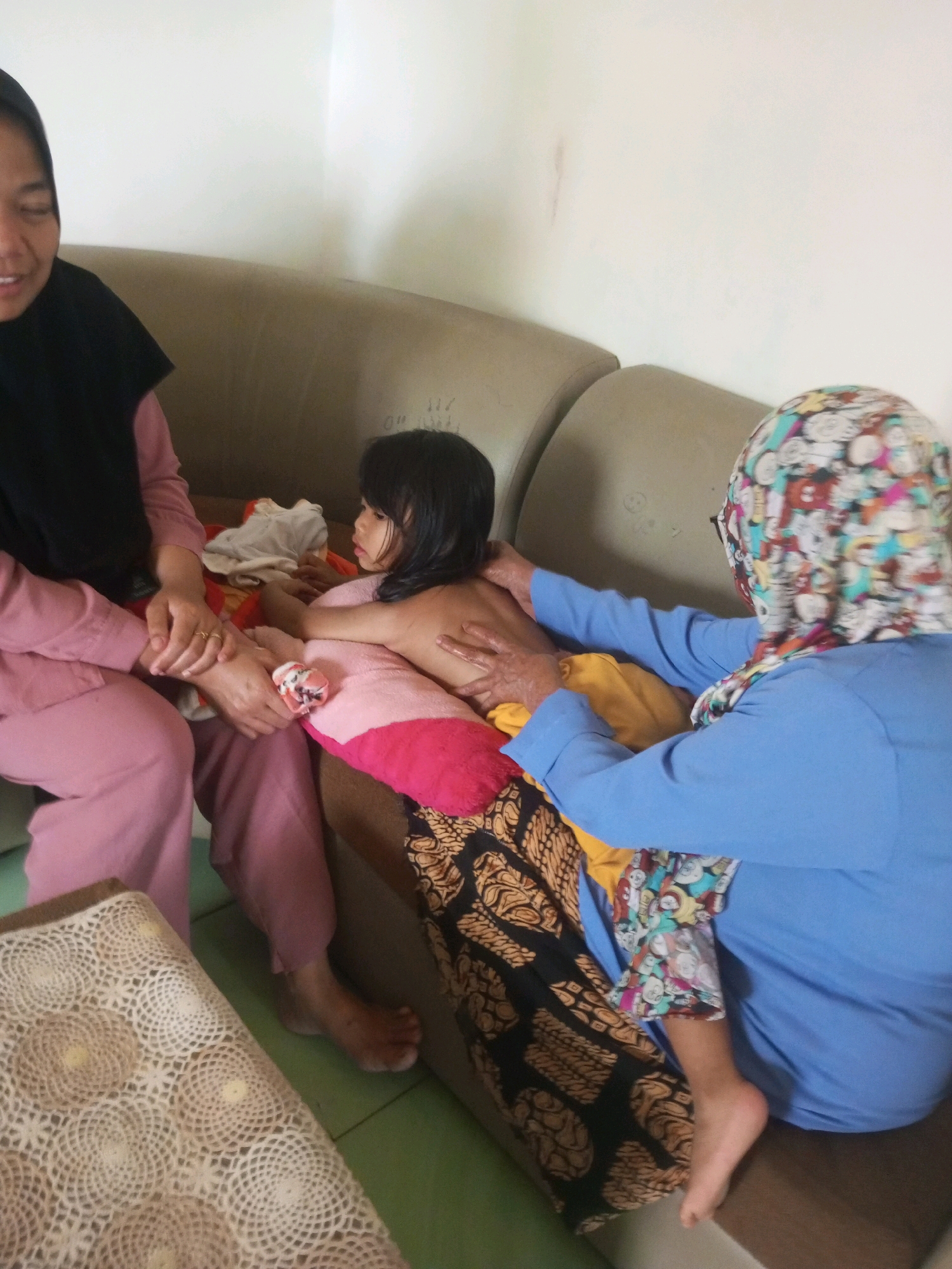 In Krasak, Indonesia, three individuals are depicted on a sofa. A woman wearing a black hijab and pink clothing sits on the left, her hands clasped over her lap. To her right, a young girl is being massaged on her back by an older woman. This woman, wearing a blue shirt and a colorful patterned headscarf, is seated behind the girl, with her hands actively working on the child's back. The girl is leaning forward, her back exposed, and she is wearing a pink garment. Another woman's arms can be seen around the girl's shoulders, implying her presence or involvement in holding the child. The sofa is upholstered in a neutral color. In the foreground, a small table with a white crocheted cover is visible. The wall behind the sofa is plain and light-colored.