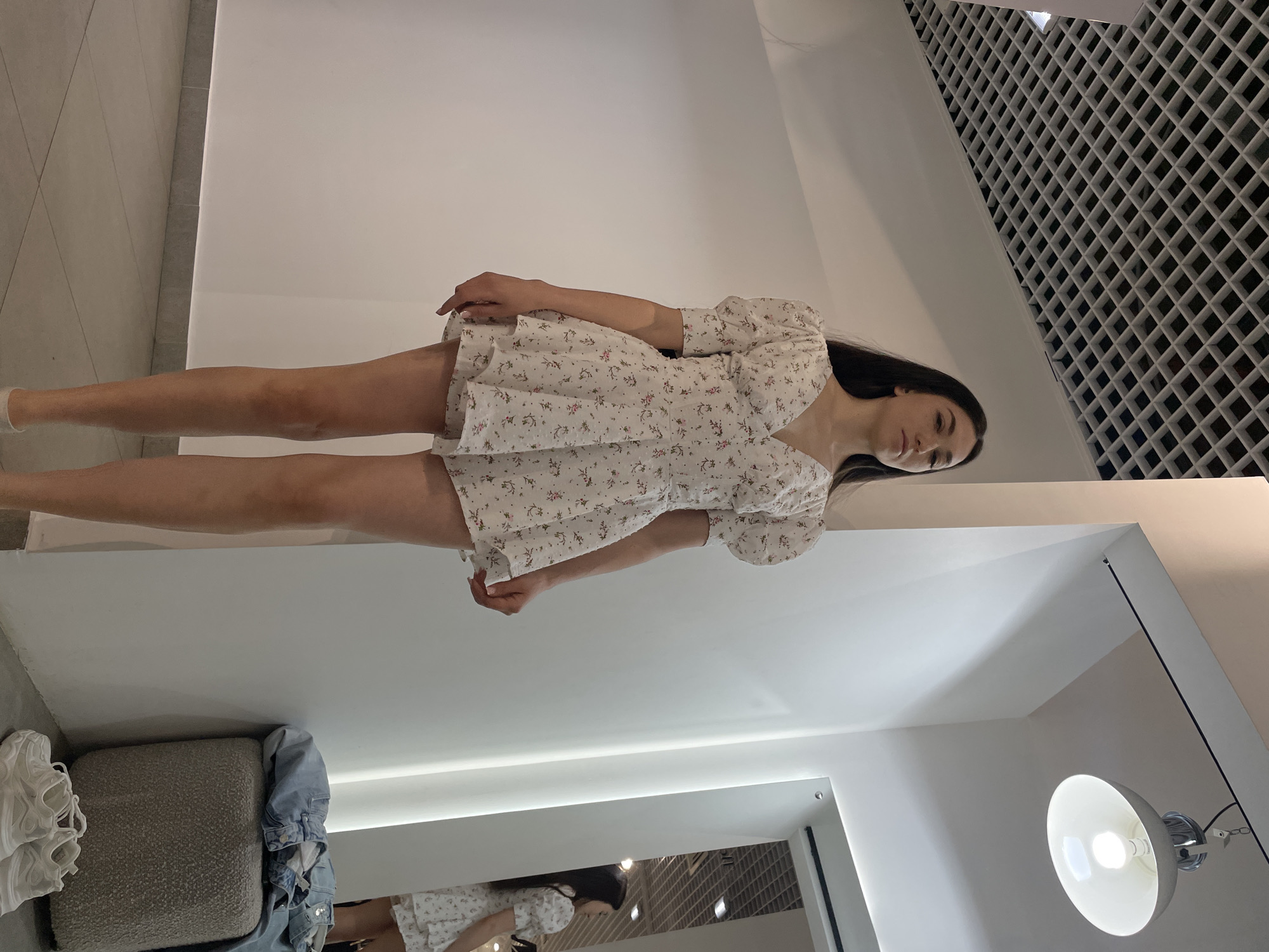 A woman with long dark hair is wearing a short, white, floral-patterned dress. She is standing in what appears to be a dressing room with white walls and a grid-patterned ceiling. The floor is covered in grey carpet, and a grey ottoman is visible in the foreground, along with a white sneaker and a denim jacket. A mirror reflects another person wearing a similar dress. A light fixture is attached to the ceiling in the bottom right corner of the image. The image was taken indoors. The exact location in Kazan, Russia, is not discernible from the image.