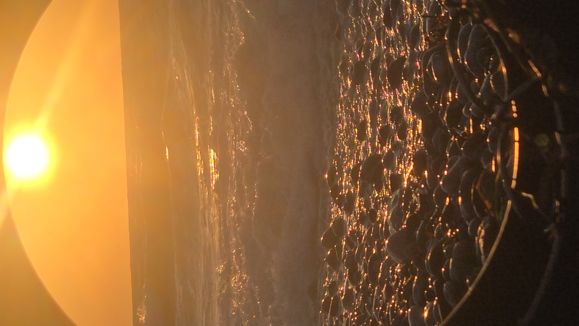 The image depicts a rocky shore at sunset, with the sun appearing as a bright, white disc on the horizon, casting a warm, orange glow over the scene. The sun's rays reflect off the gentle waves of the ocean, creating shimmering highlights on the water's surface and on the wet pebbles of the shore. The foreground is dominated by a collection of dark, smooth stones, some of which are wet and glisten with reflected light. In the upper portion of the image, a curved, dark shape is visible, framing the sun and the ocean. This framing element could be part of a structure or natural formation. The overall atmosphere is serene and tranquil, suggesting a natural coastal environment. The location is identified as Soroni, Greece.