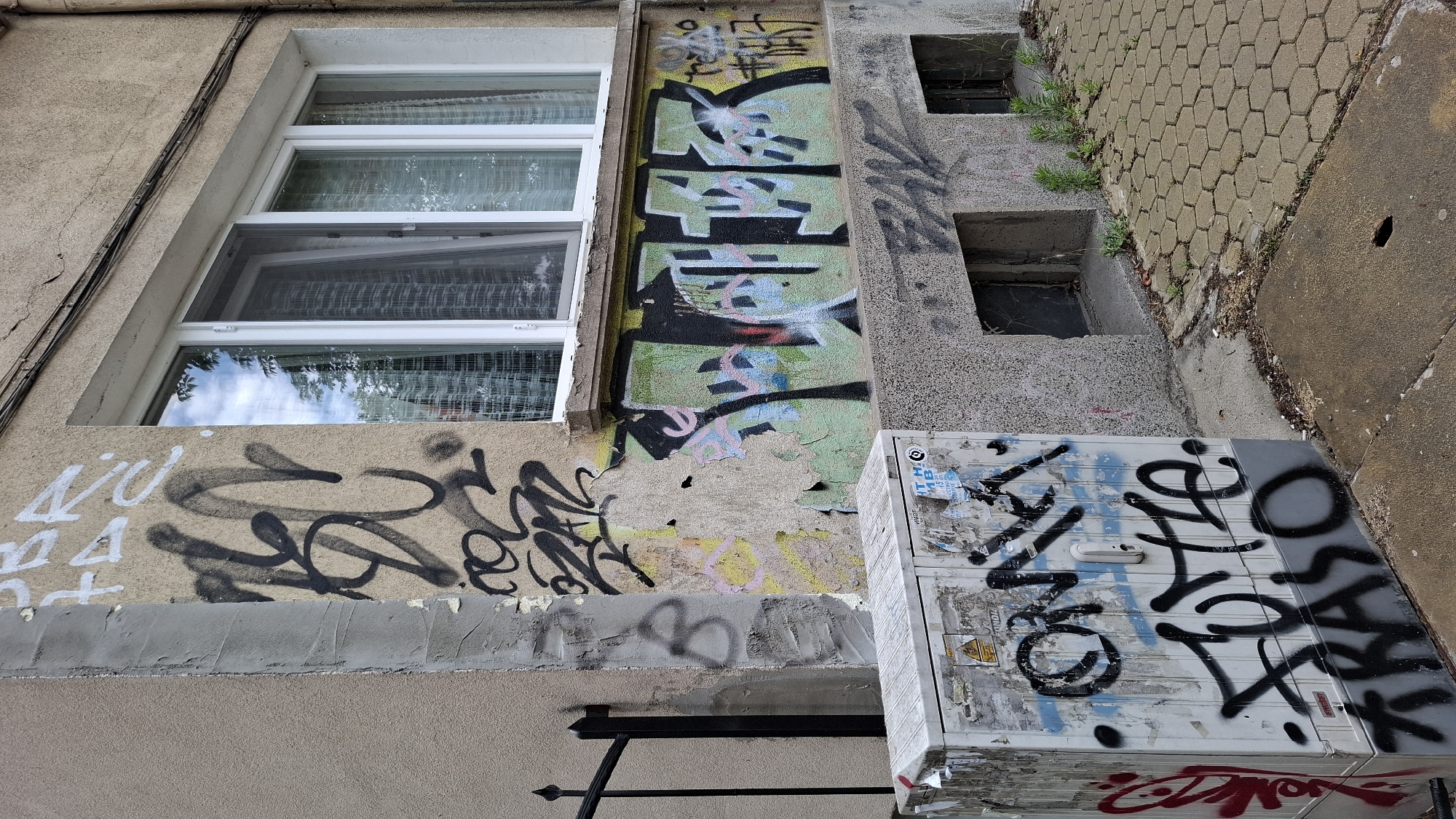 The image depicts the exterior of a building in Sofia, Bulgaria, characterized by significant graffiti. The building has a tan-colored facade with a visible crack above a white-framed window unit featuring sheer curtains. The lower portion of the building displays a band of graffiti in black, pink, and green spray paint, with the prominent green graffiti appearing to spell out "FLOK" or a similar word in a bubble letter style. To the left of this, on a slightly damaged and discolored section of the wall, are further graffiti tags in black and white. In the foreground, a white electrical cabinet is covered in multiple layers of graffiti, including tags in black and red spray paint. Notable tags on the cabinet include "OMIK," "FUTE," and "KRASO." The ground surface consists of concrete and hexagonal pavers, with scattered debris and small plants growing in crevices. A black metal fence is partially visible on the far left. The scene suggests an urban environment with signs of neglect and vandalism.