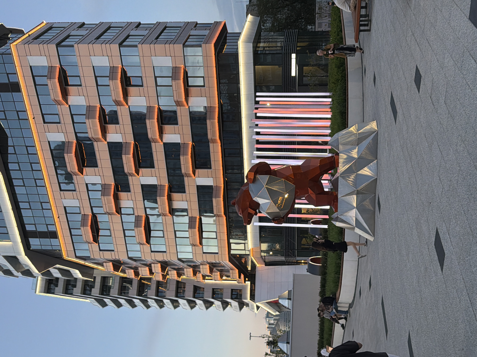 The image depicts a modern urban setting in Yekaterinburg, Russia. A tall, multi-story building with a unique facade dominates the left side of the frame. The building features alternating blocks of glass and a copper-toned material, with architectural elements that jut out around the windows. To the right of the building, there is a sculpture of a bear made of geometric, faceted shapes, primarily in reddish-brown and silver tones.
In the background, behind the bear sculpture, there is a set of horizontal light bars emitting a pinkish-red glow. A few individuals can be seen walking or standing in the vicinity. The ground is paved with light gray tiles, with a scattered pattern of dark squares. The sky is a pale blue, suggesting either early morning or late evening lighting conditions.