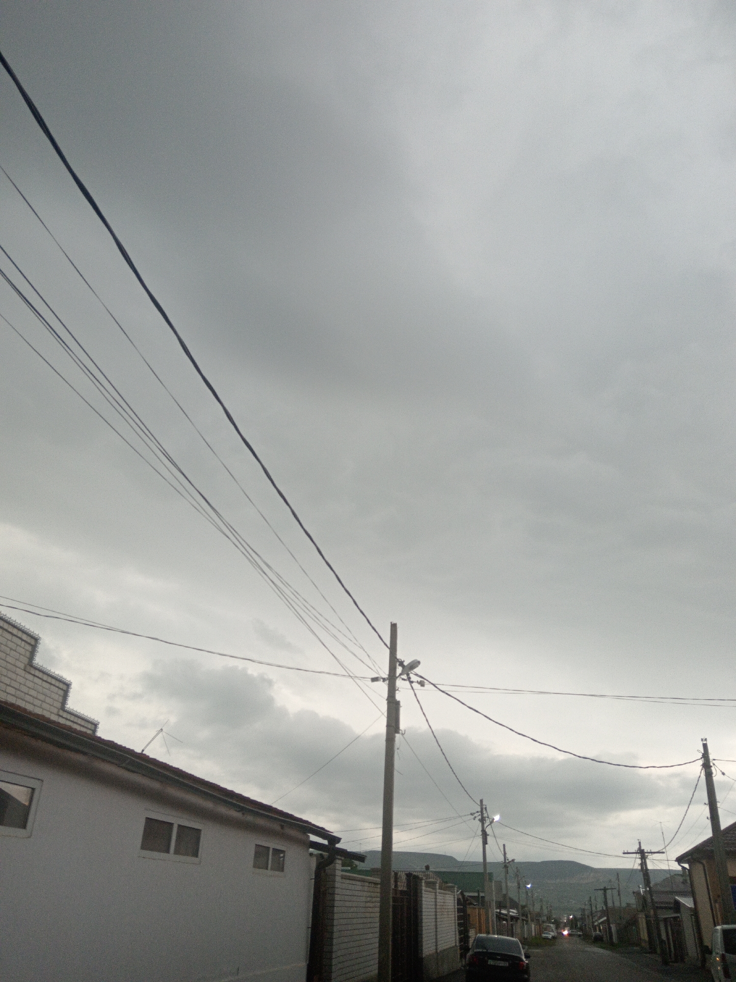This media file shows a street scene in Uchkeken, Russia. The image is dominated by a cloudy, overcast sky. Multiple power lines stretch across the frame, suspended from poles that line the street. On the left side of the image, a white building with several windows is visible, partially obscured by the power lines. Further down the street, more buildings and fences can be seen, suggesting a residential or rural area. A few cars are parked or driving on the street, including a dark-colored car in the foreground. In the distance, hills or mountains are faintly visible through the haze. The overall mood is somber, likely due to the cloudy weather. The time of day is difficult to determine precisely, but the lighting suggests it could be late afternoon or early evening. No text is discernible in the image.