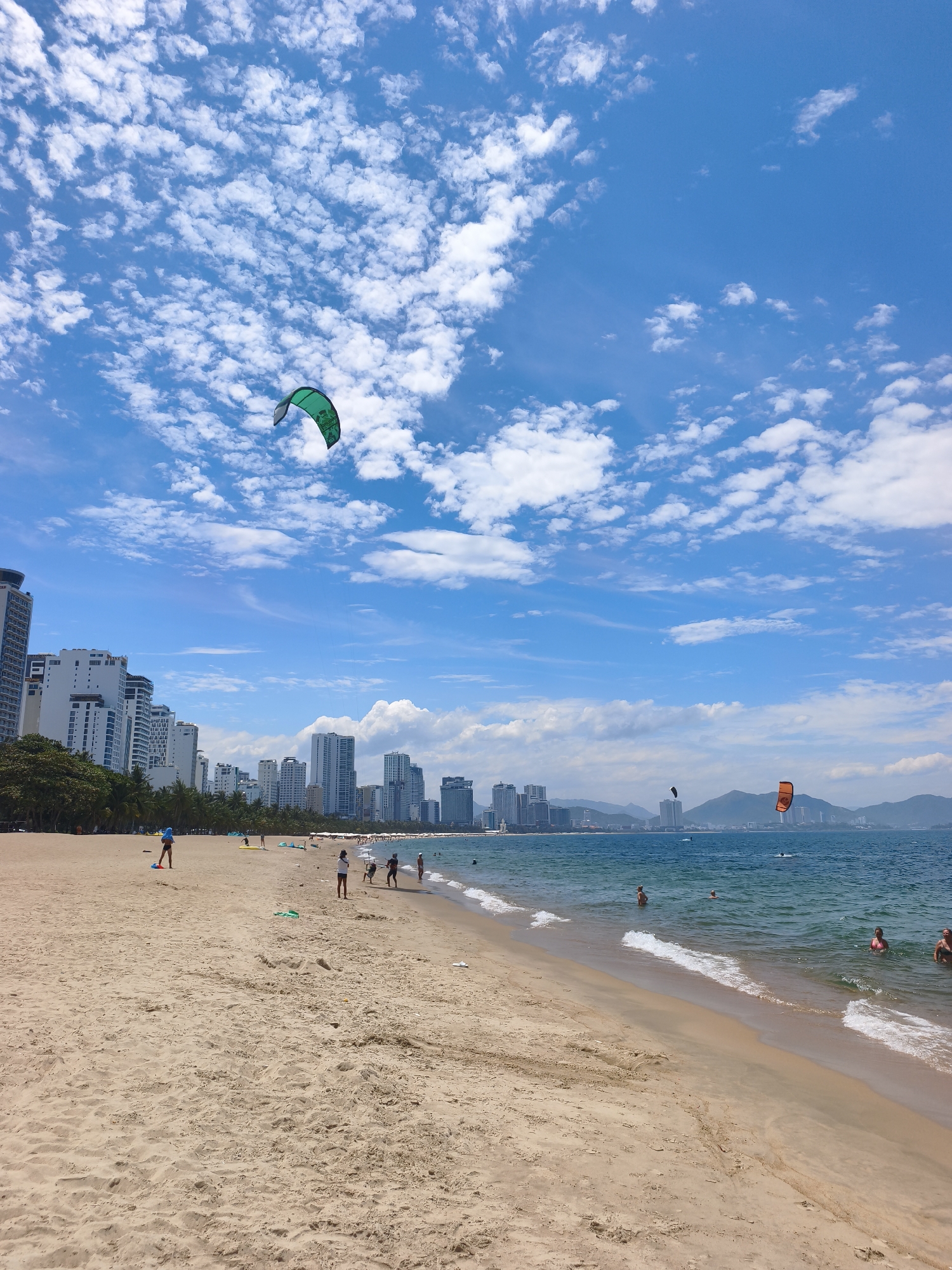 This is a medium-shot, eye-level, outdoor, daytime photograph of a beach in Nha Trang, Vietnam. The sandy beach occupies the foreground, with a line of palm trees and a cityscape of tall buildings in the background. The ocean is visible on the right side of the image, with waves gently lapping the shore. A few people are on the beach and in the water. In the sky, there are many white, fluffy clouds. A green kite is visible in the upper left portion of the sky. Two more kites, one orange and one black, are visible in the distance over the ocean. The general setting is a sunny day with a clear blue sky.