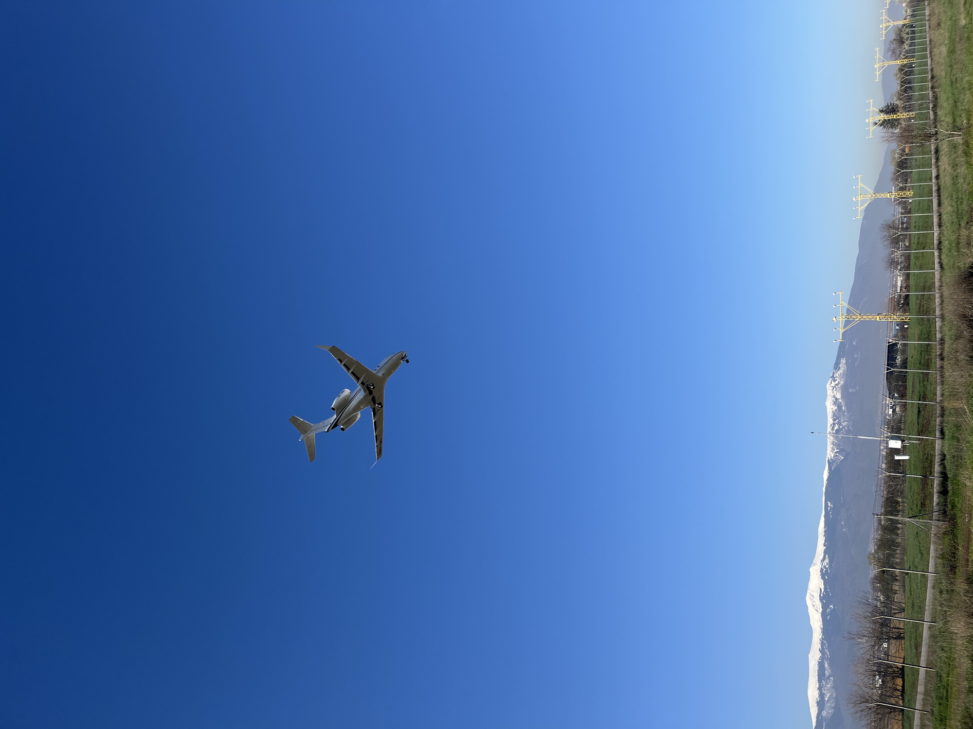 A low-angle shot captures a white aircraft in flight against a bright blue sky. On the right side of the image, there's a landscape featuring mountains with visible snow on the peaks, and a grassy area with what appears to be infrastructure such as power lines. The location is undetermined, no information about the location in the city of Krivina, Bulgaria can be found in the image.