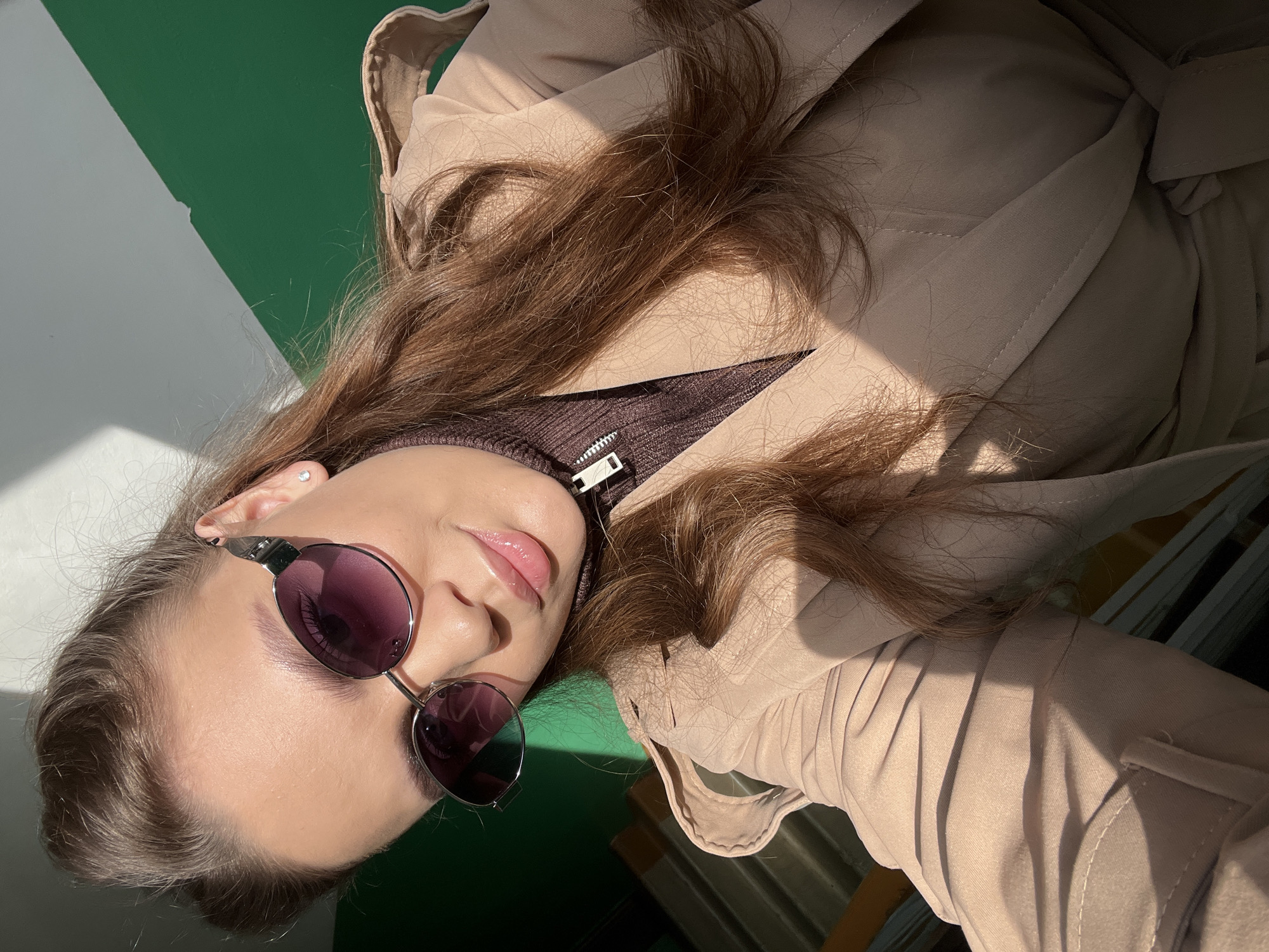 A young woman with light skin, long brown hair, and wearing round sunglasses is the primary subject of this portrait. She has a brown turtleneck sweater underneath a tan trench coat. She is wearing an earring in her left ear.
The background consists of a split white and green surface. The lighting suggests a sunny day.
The image does not contain enough information to determine its location within Chaikovsky, Russia.