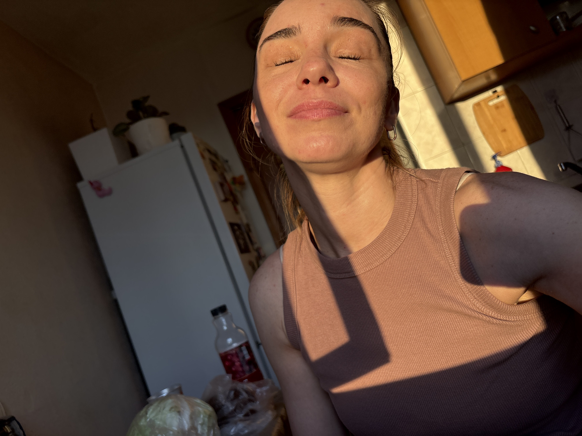 A woman is standing in what appears to be a kitchen, with sunlight streaming in from the right, casting a strong shadow across her. She is facing forward with her eyes closed, a serene expression on her face. She is wearing a pink ribbed tank top. In the background, to her left, is a white refrigerator with a plant on top. To the right, a wooden countertop is visible, along with kitchen cabinets. A bottle of red liquid and some plastic-wrapped items are on the floor in front of the refrigerator. The overall setting suggests a casual, domestic environment bathed in natural light. The location is identified as Unknown, Russia.