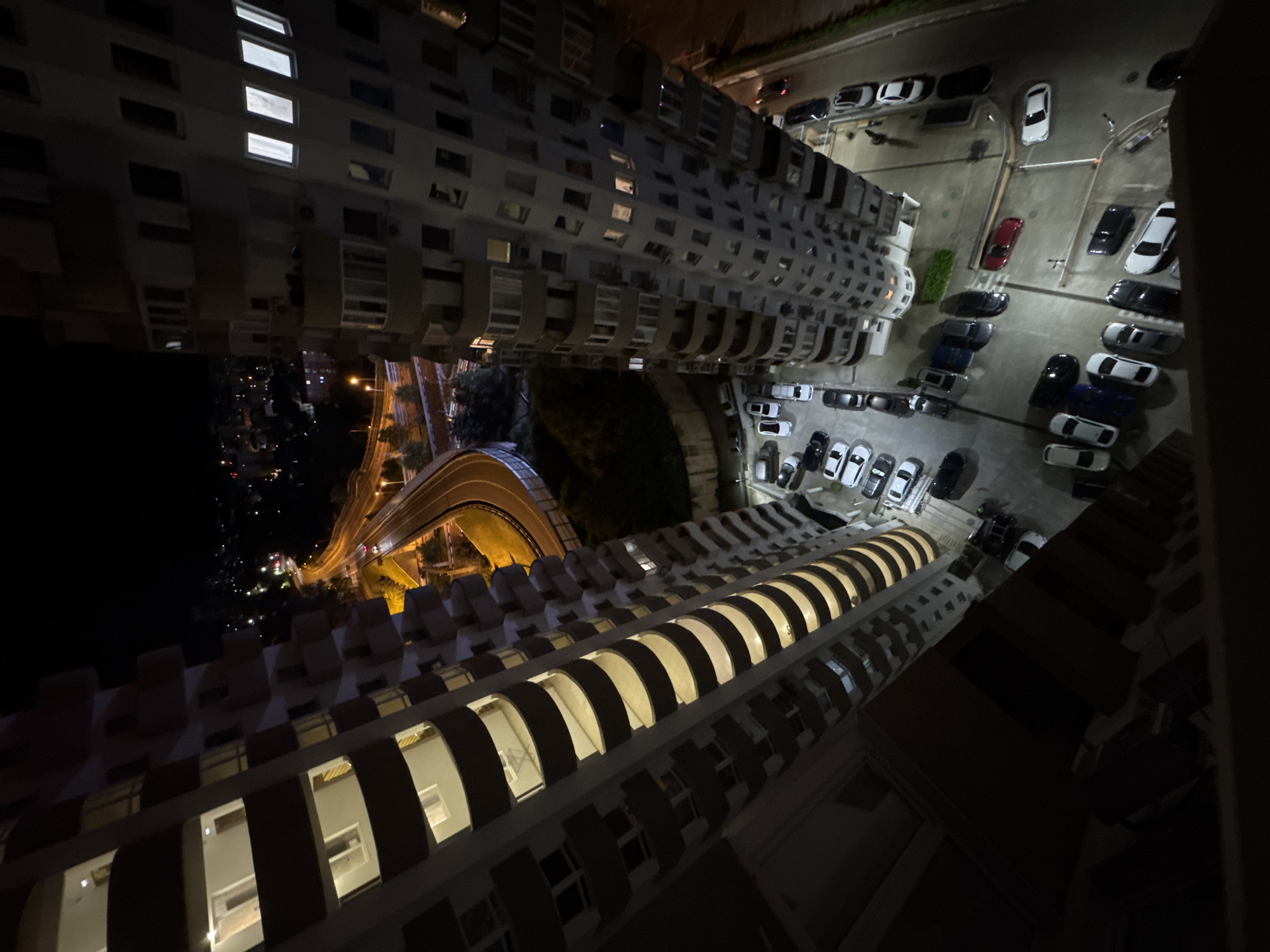 This high-angle, downward-looking night photograph captures a dense urban environment within the city of Sochi, Russia, likely taken from a high floor of a residential building. The scene is dominated by two tall, multi-story residential buildings flanking a complex of roads and parking lots.
On the left, a light-colored building with numerous dark, curvilinear balconies extends vertically through the frame. Many of its apartments are illuminated from within, casting a warm light onto the balconies. A small white, possibly collapsible, table is visible on one of the mid-level balconies.
On the right, another multi-story residential building with a lighter facade features numerous rectangular windows, some of which are lit. Several external air conditioning units are visible on its facade.
Between these two buildings, and beneath an elevated road section, are multiple asphalt parking lots. These lots are filled with numerous parked passenger vehicles, primarily sedans and SUVs, in various colors including dark hues, white, silver, and one prominent red car. Ground markings are faintly discernible in the parking areas. In the upper right section of the parking area, two indistinct human figures appear to be walking.
Above the ground-level parking, an elevated asphalt highway or arterial road curves sharply from the left toward the right, illuminated by yellow-orange streetlights. A lower road section is visible beneath and to the left of the elevated portion. Guardrails delineate parts of the road infrastructure.
The background reveals a vast expanse of distant city lights scattered across hilly or mountainous terrain, receding into the darkness under a clear night sky. Patches of dark foliage and landscaping are present around the parking areas and roads. The foreground is framed by the dark, out-of-focus edge of a structure, presumably the balcony or railing from which the photograph was captured.