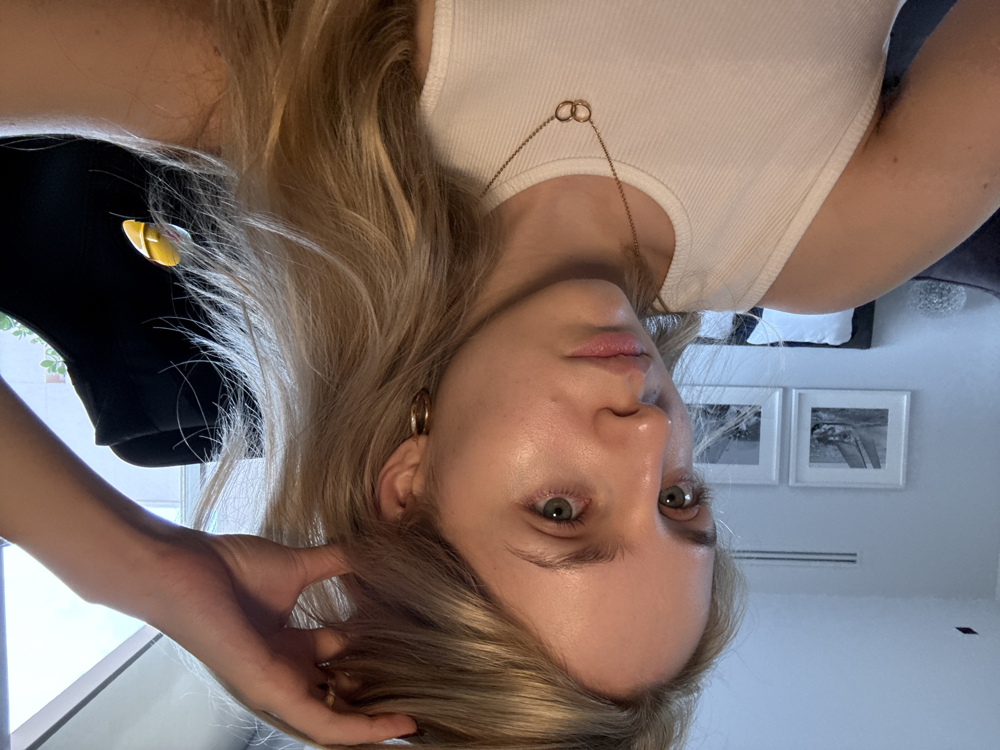 A woman with blonde hair, fair skin, and green eyes is looking at the camera. She is wearing a white tank top and a gold necklace with interlocking circles. She has a gold hoop earring on her left ear. Her left hand is in her hair.
In the background, there is a black garment hanging near a window. Behind the woman, there are two framed black and white photographs on a white wall. The location appears to be indoors, possibly a home. Based on the style and architecture, the location could be Mexico City, Mexico.