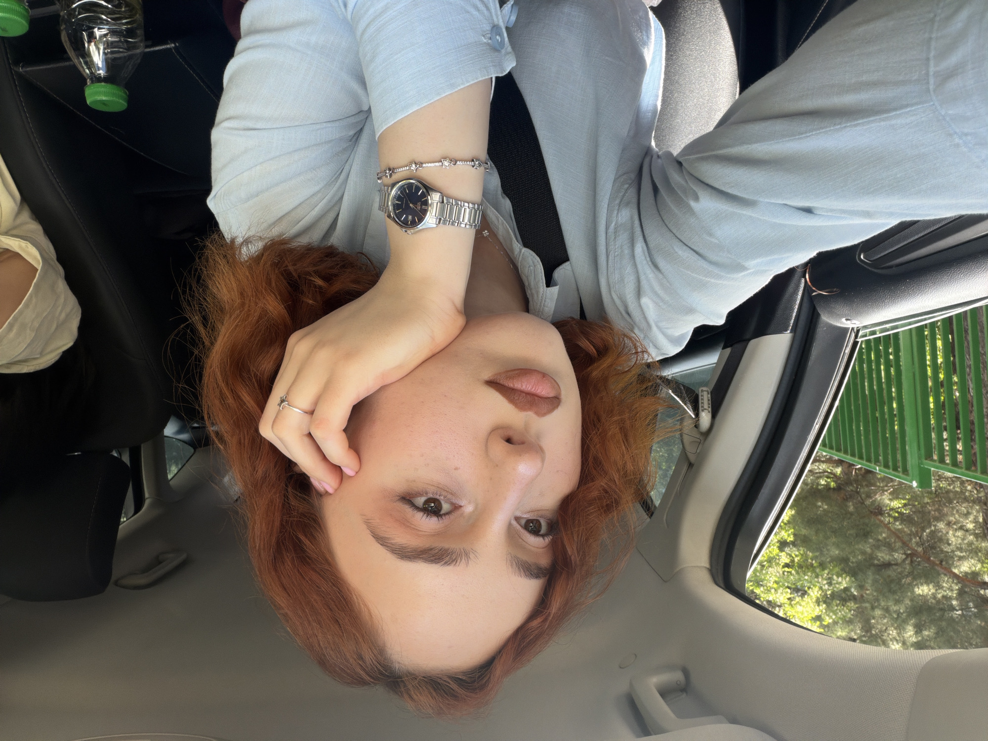 A woman with red hair is photographed inside a car. She is wearing a light blue button-down shirt and a metal watch on her left wrist. Her face is partially obscured by her left hand, which has a ring on the ring finger. The background visible through the car window includes greenery and a green fence, potentially indicating a park or wooded area. Given the prompt's reference to Tbilisi, Georgia, the location is presumed to be within that city. A water bottle is visible in the upper left corner. The photograph has been rotated, placing the woman's head at the bottom of the frame.