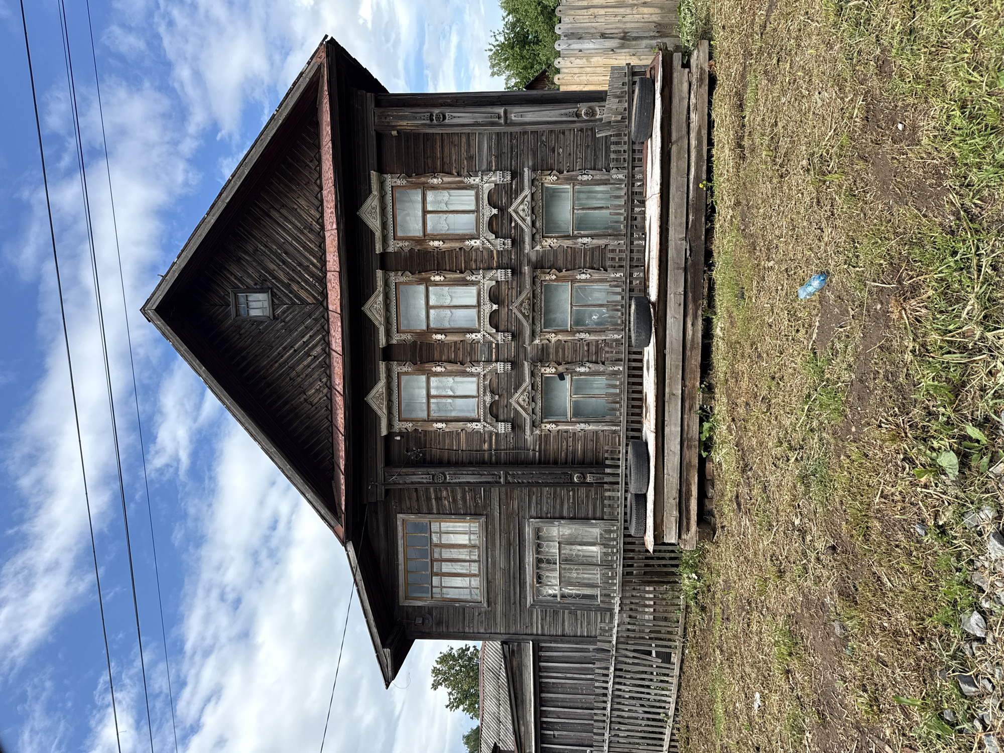 The image, correctly oriented, depicts a weathered, traditional wooden house in Votkinsk, Russia, under a bright blue sky dotted with white clouds. The house, constructed from dark timber, showcases distinctive Russian architectural elements, particularly the intricately carved wooden window frames, known as "nalichniki." These elaborate frames, with their geometric and ornamental patterns, adorn several windows on the house's main facade.
The building features a prominent, steeply pitched roof with dark shingles and a small, square attic window centered on the gable end. The facade displays multiple windows across two visible stories, each surrounded by decorative carvings. Some windows show faint white curtains, while others reflect the sky.
To the right of what appears to be a porch or balcony area, two old tires are visible, potentially used as planters or for storage. A simple wooden fence extends along the front of the property, separating the house from a patch of dry, sparse grass and dirt. A discarded blue plastic bottle lies on the ground near the fence. In the background, beyond the house and fences, green tree foliage is visible. Utility lines traverse the upper left portion of the sky.
The scene captures a sunny daytime moment in a quiet, likely rural or semi-rural setting. There are no people present, and no specific activities are taking place. The weathered wood and traditional design elements convey a sense of age and local cultural heritage. No visible text is present in the image.