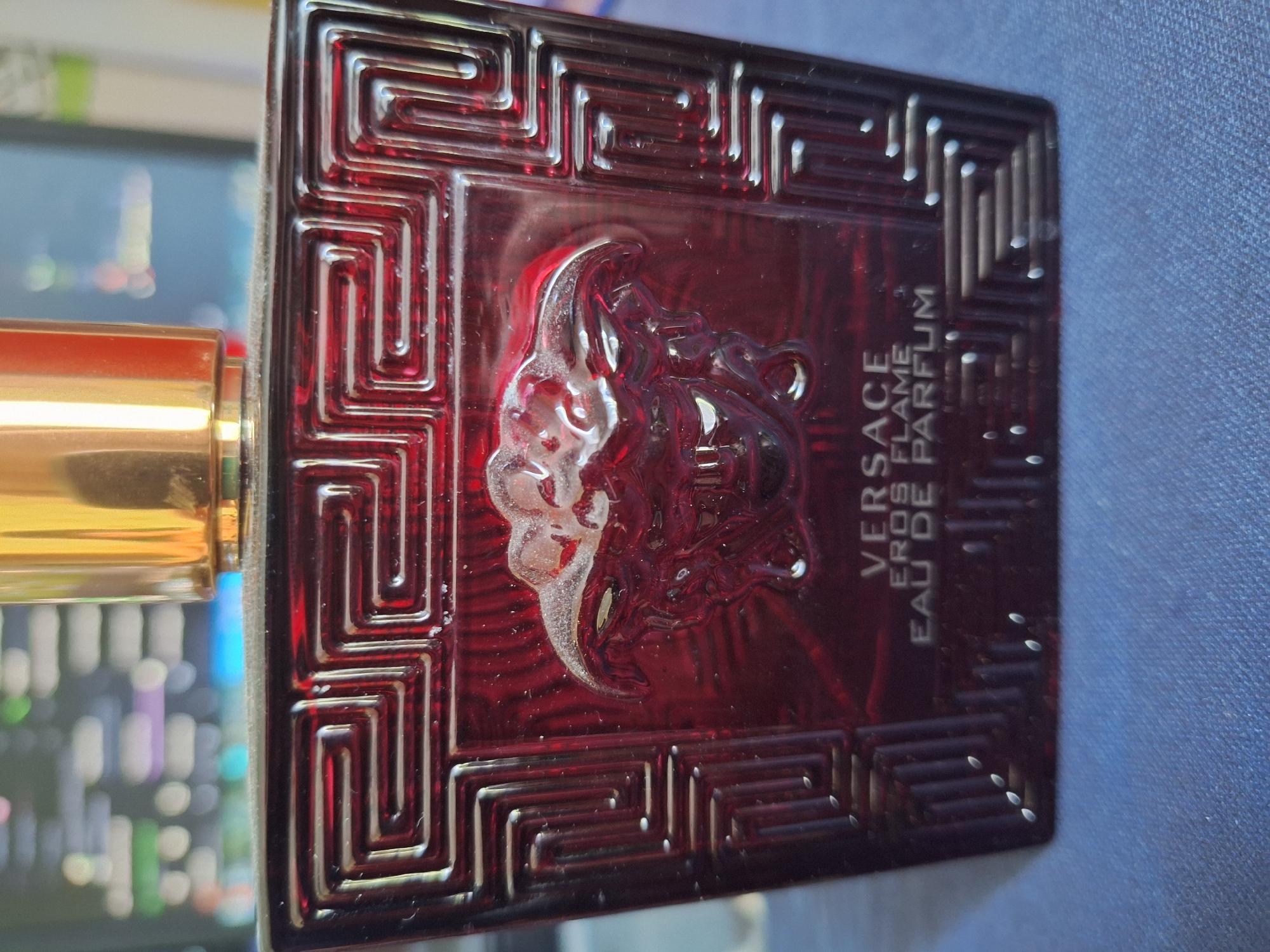 A close-up shot focuses on the front of a red Versace Eros Flame Eau de Parfum bottle. The bottle has a distinctive Greek key pattern etched into its surface and a raised Medusa head emblem in the center. The cap of the bottle is a shiny gold color. The background is blurred, showing what appears to be a computer monitor with various colored lights and text. The bottle is resting on a textured blue surface, possibly a table or fabric. The image's lighting highlights the details of the bottle, casting subtle shadows. No specific location in Varna, Bulgaria, is indicated.