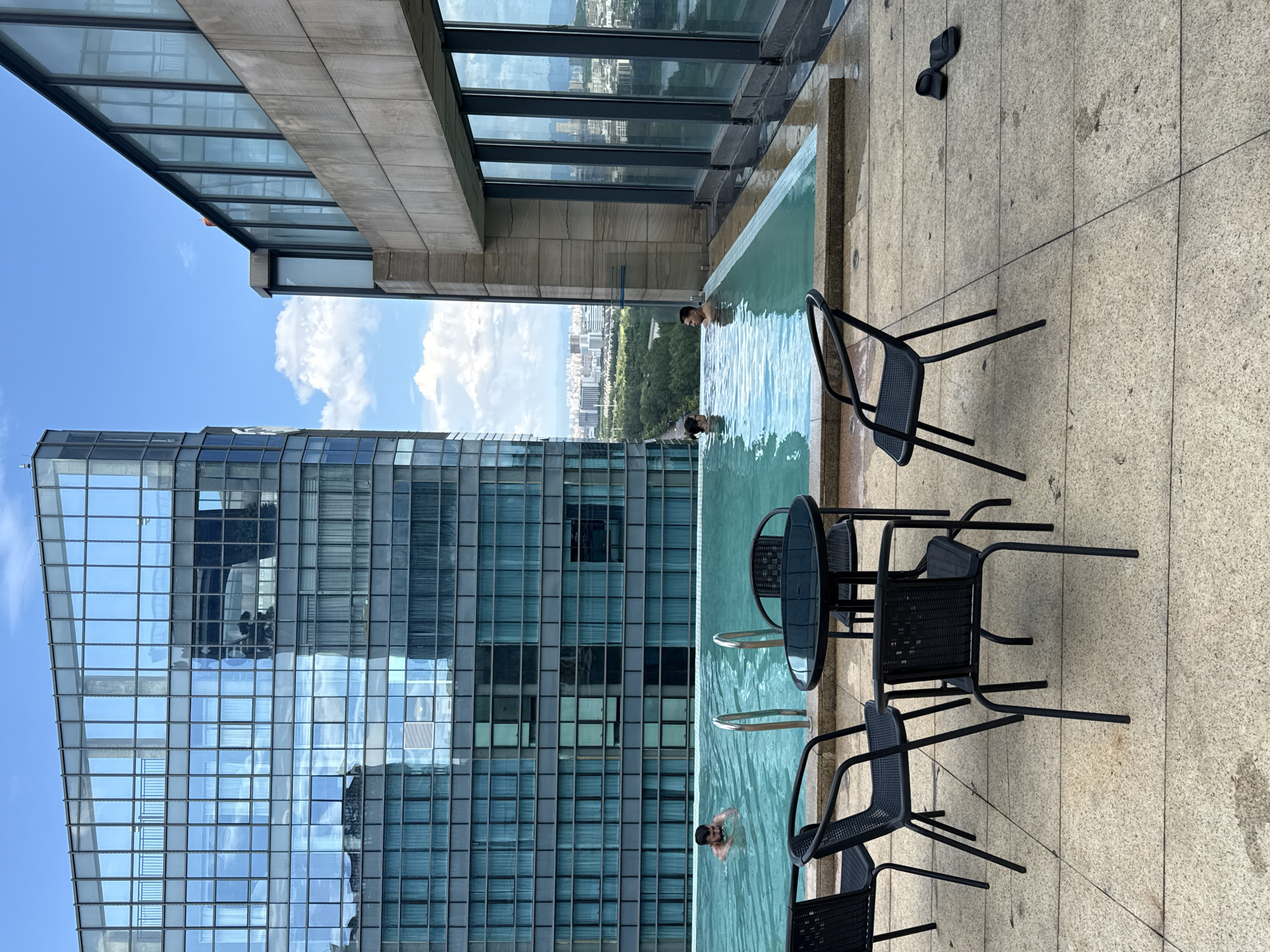 A rooftop swimming pool is pictured with a city view in Yiwu, China. Two men are in the pool, with one man partially submerged up to his chest and the other man standing with his head and shoulders visible. In the foreground, black metal chairs and a table are arranged on a paved area next to the pool. A tall, modern building with a glass facade dominates the left side of the image, and another building with a stone facade is on the right. The sky is blue with scattered clouds. The distant cityscape shows a mix of buildings and greenery.
