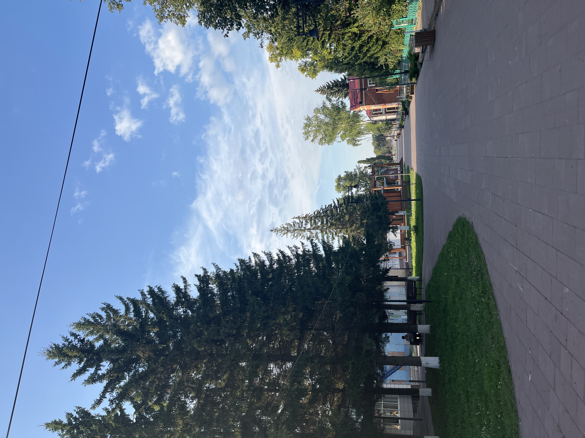 The image depicts a wide, paved pedestrian walkway lined with trees and buildings. On the left side of the walkway, there are several tall evergreen trees and a grassy area with benches. A person is seated on one of the benches, facing away from the camera. Behind the trees, there are low-rise buildings with windows.
The walkway extends into the distance, where more trees and buildings can be seen. On the right side of the walkway, there are more trees, some lampposts, and a green fence. The sky above is blue with scattered white clouds.
The scene appears to be a public park or a street with a pedestrian area, possibly in a town or city. The overall impression is one of a calm and pleasant outdoor environment.
The provided location of Novye Turbasly, Russia, suggests a specific geographic setting for this scene.
