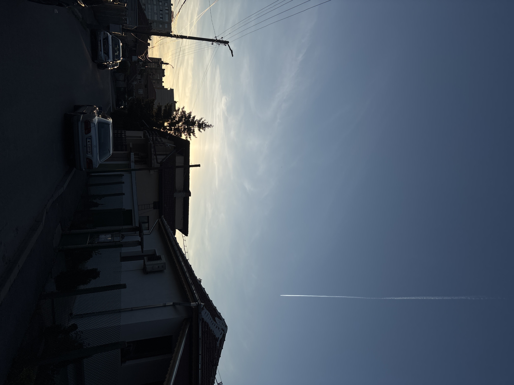 The image shows a street scene in Lom, Bulgaria, under a bright, clear sky with a contrail. Two cars are parked on the left side of the street. One is a silver sedan, and another is a dark-colored SUV. On the left side of the street is a building with a gray exterior and a red roof. The sky is clear, with a jet contrail visible. Overhead electrical wires span the width of the street.