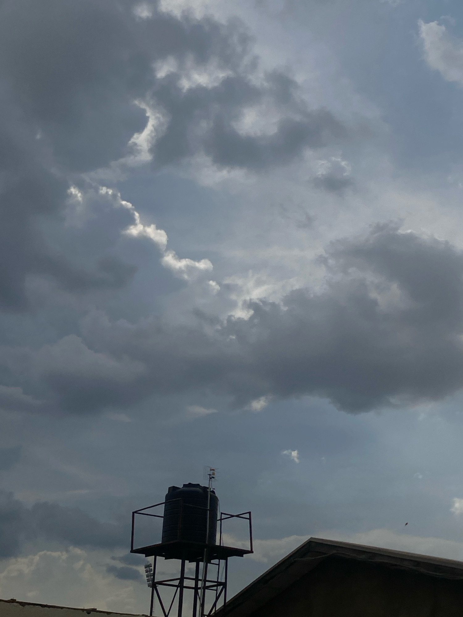 The image depicts a water tank perched atop a metal structure, situated on a rooftop in Dadi Riba, Nigeria. The sky above is heavily clouded, with dark, heavy storm clouds dominating the scene, hinting at impending rain. Sunlight occasionally breaks through the clouds, creating pockets of brighter light. A portion of a building's roof is visible in the foreground, and a small antenna is mounted on top of the water tank structure. No people are visible in the frame.