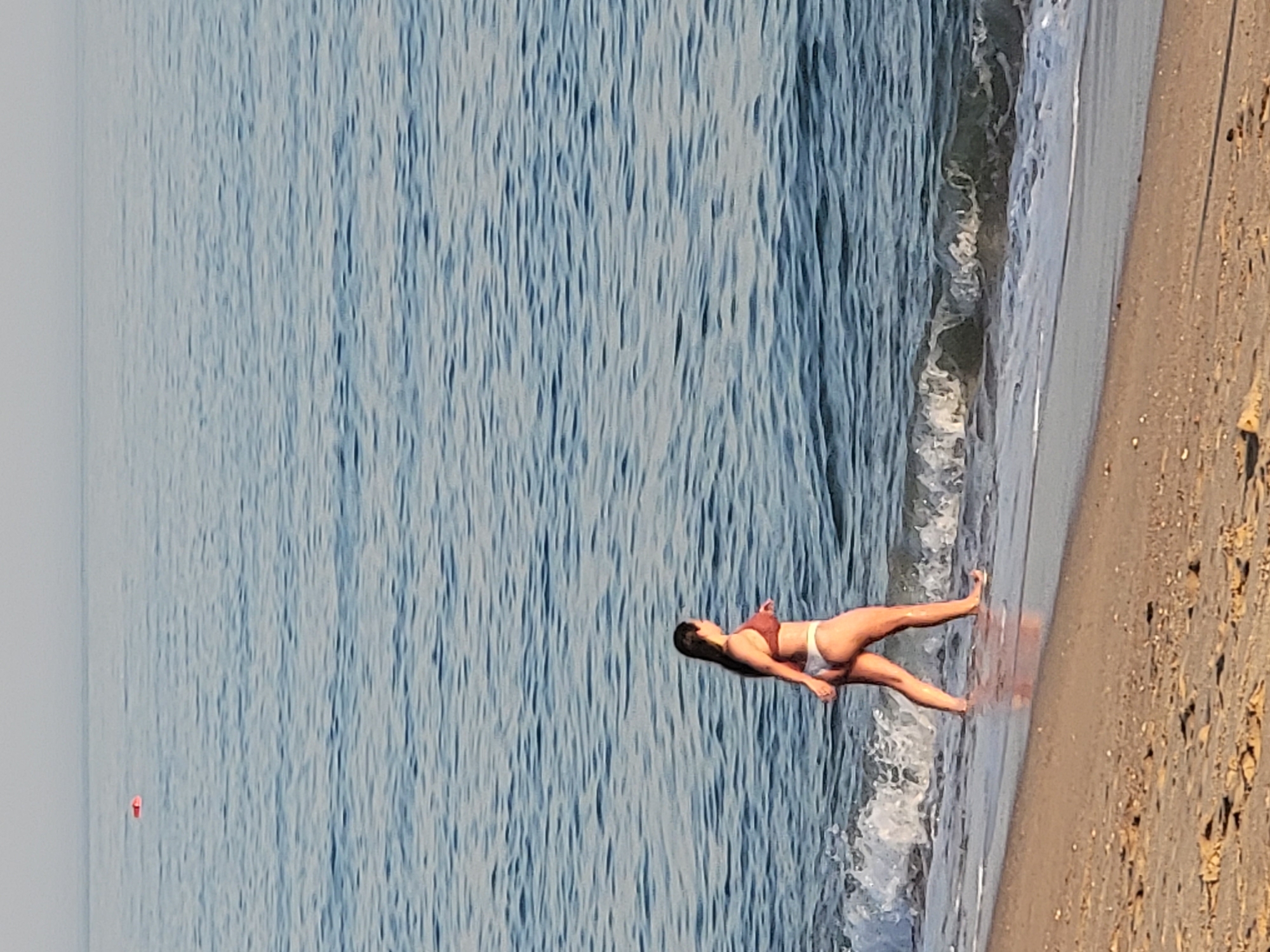 A full-body shot shows a woman walking along the shoreline of a sandy beach in Quarteira, Portugal. The woman is facing right and walking into the water, her body is slightly turned to the left. She is wearing a brown bikini and her long, dark hair is down. The ocean has light blue, rippling water, with small waves crashing on the shore. The sky is a pale blue, suggesting early morning or late afternoon light. In the distance, there is a small red object floating on the water. The beach is sandy with some small debris scattered on it.
