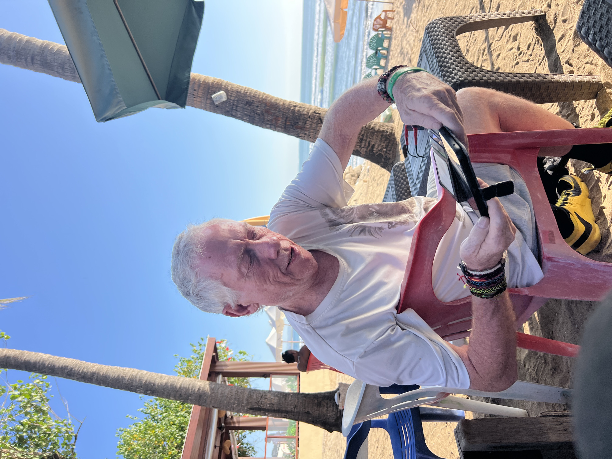 An elderly Caucasian male with gray hair and weathered skin sits on a red plastic chair on a sandy beach. He is wearing a white t-shirt, light-colored shorts, and yellow and black athletic shoes. He has multiple colorful bracelets on his left wrist. The man is looking down at a black object, possibly a phone or small tablet, in his hands. His right hand is reaching towards the object.
In the background, palm trees line the shore, and the ocean with gentle waves is visible. Beach chairs and umbrellas are scattered along the sand. A building with a brown roof and wooden structures is partially visible to the left. Another person, wearing a dark shirt, sits on a red chair in the distance. The scene appears to be a beach in Kuta, Indonesia, indicated by the tropical vegetation, ocean, and beach atmosphere. The lighting suggests it is daytime with clear skies.