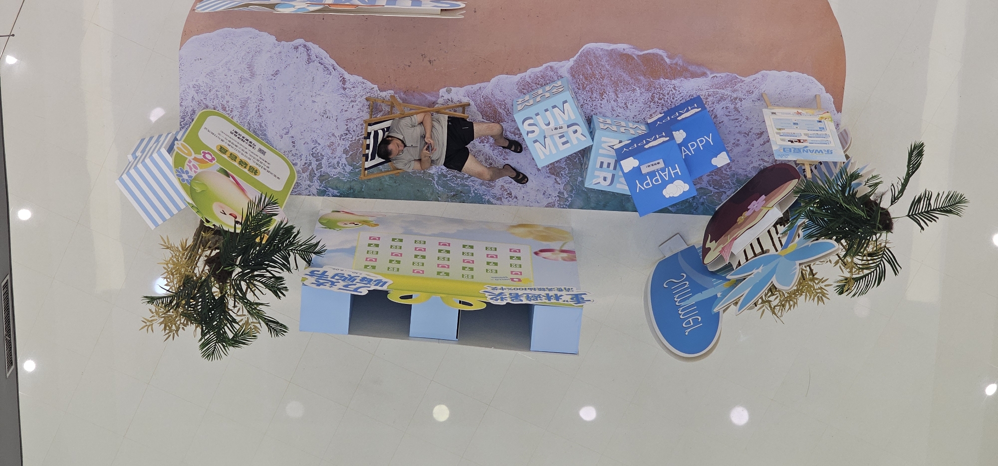 An overhead shot captures a man sitting in a beach chair on a decorative beach scene, which appears to be a promotional display. The man, wearing a grey polo shirt and black shorts, is looking down at his phone. The beach display features a light pink sand-colored floor with white wave-like foam decorations. Scattered around the display are blue cubes with white text, including "SUMMER" and "HAPPY." The scene also includes artificial palm trees and floral decorations. To the left of the man, there is a large display board with Chinese text and a cartoon image of a green apple, likely part of the promotional setup. The floor beneath the display is a light-colored tile. The location is identified as Shapingba District, China.
