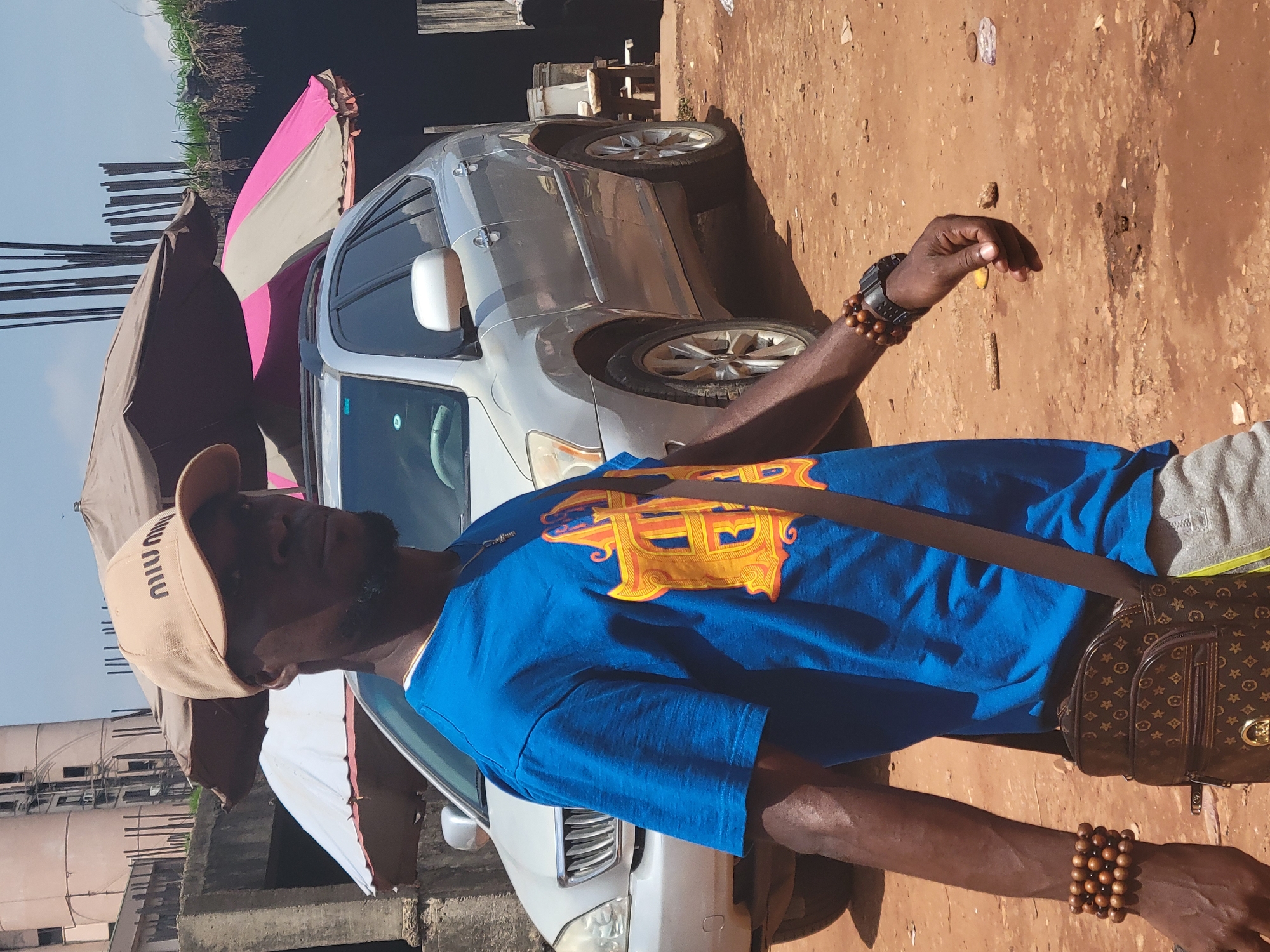 The image depicts a dark-skinned man standing outdoors in what appears to be a street or informal market setting in Abuja, Nigeria, during daylight hours. He is wearing a beige baseball cap with the word "MIUMIU" printed on it, a bright blue t-shirt featuring a large, ornate yellow and orange monogram graphic (possibly interlocking letters like "H" and "J" or "R"), and grey shorts. He sports a black watch on his left wrist and multiple wooden beaded bracelets on both wrists. A dark brown shoulder bag with a monogram pattern, characteristic of Louis Vuitton designs, hangs across his body. His right hand is slightly raised with fingers curled, and he has a short beard.
Behind the man, a silver car, possibly a Toyota sedan, is parked. To the left and right of the man are large canopies or umbrellas; one is dark brown, and another is grey and pink. The ground is a reddish-brown dirt path, scattered with small stones and debris. In the background, under a clear blue sky, there are glimpses of structures with exposed rebar and an area with dark water or mud and sparse vegetation, suggesting an urban or semi-urban environment. The scene is brightly lit, indicating sunny weather.