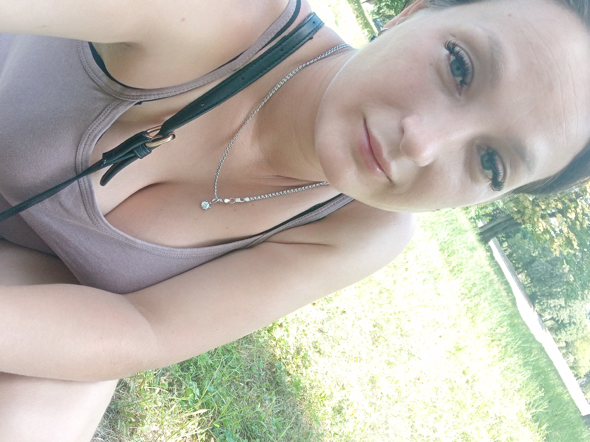 In a sun-drenched park in Chuhuiv, Ukraine, a woman with striking blue eyes and long, dark eyelashes gazes directly at the camera. She is dressed in a simple, taupe-colored tank top, and a silver chain with a diamond pendant rests on her collarbone. Her dark hair is pulled back, and she wears subtle makeup, highlighting her features. The background suggests a relaxed outdoor setting, with lush green grass and trees visible, indicating a pleasant day. The woman appears to be seated, with a portion of her shoulder and arm visible in the frame, and a dark bag strap crossing her chest. The overall impression is of a casual, candid moment captured in a public green space.