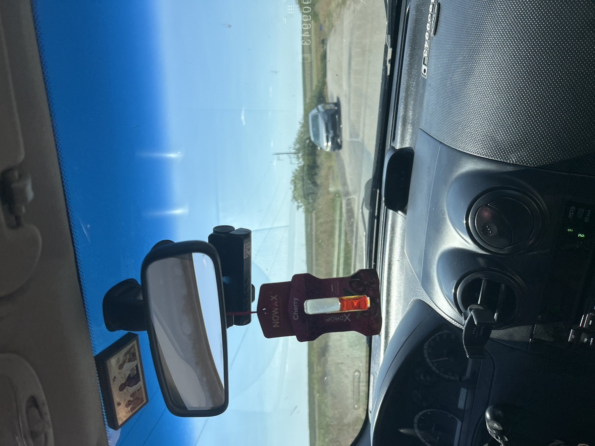 The image shows the interior of a car, looking out through the windshield. In the foreground, the dashboard of the car is visible, with an air vent and part of the steering wheel. Hanging from the rearview mirror is a cherry-scented car air freshener. A small framed image, possibly religious icons, is attached to the windshield above the rearview mirror. In the middle ground, a dark-colored car is visible on a road, with dry vegetation and a utility pole on either side. The background consists of a clear blue sky with some distant clouds and fields. The scene appears to be in a rural or semi-rural area. The location is identified as Comrat, Moldova.