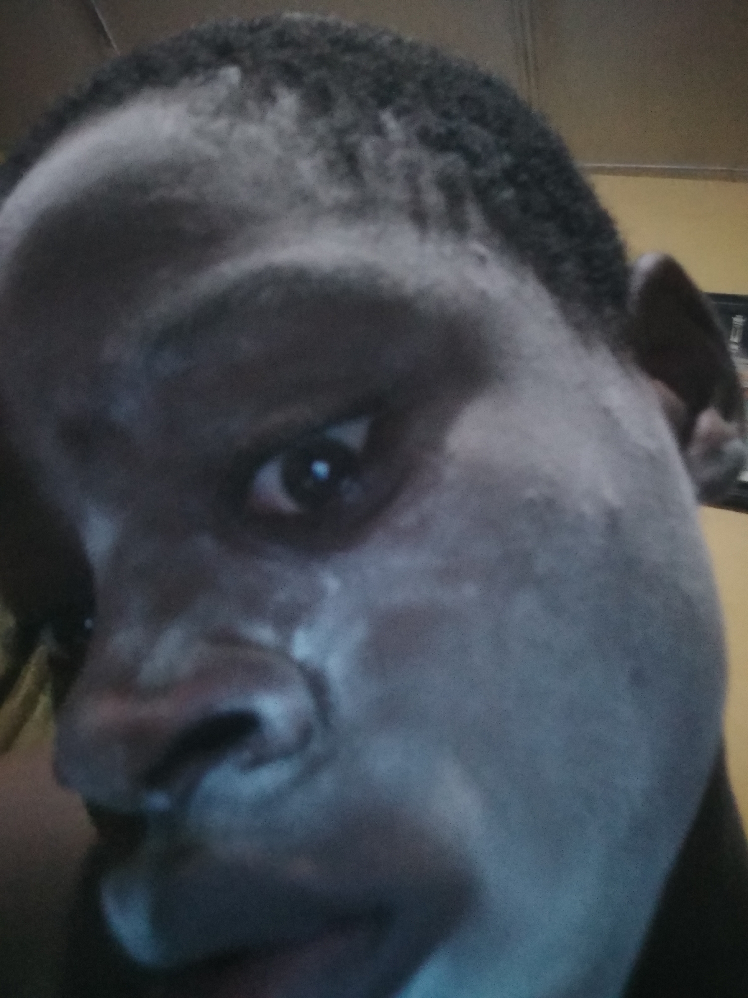 Close-up shot of a person's face with dark skin. The face is tilted towards the camera. The person has short, dark hair. There is a slight sheen on the skin, possibly due to moisture or product. The background is blurry and indistinct, suggesting the focus is on the person's face. No indication of Shangisha, Nigeria is visible.