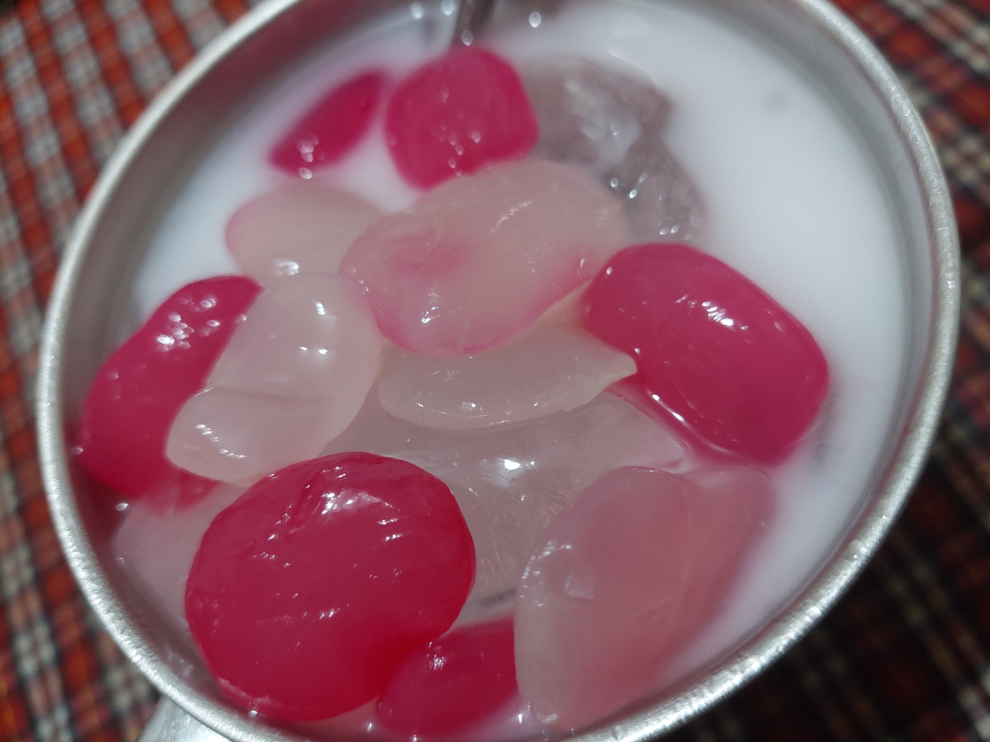 A close-up, overhead view shows a metallic bowl filled with a white liquid and several gelatinous, oval-shaped pieces. Some of the pieces are bright pink, while others are translucent white or pale pink. Ice cubes are visible beneath the pieces and the liquid. The bowl is positioned on a surface covered with a red and white checkered cloth. The scene is well-lit, with reflections visible on the surface of the gelatinous pieces and the liquid. The image does not provide specific details about the location being in Jelutung, Indonesia.