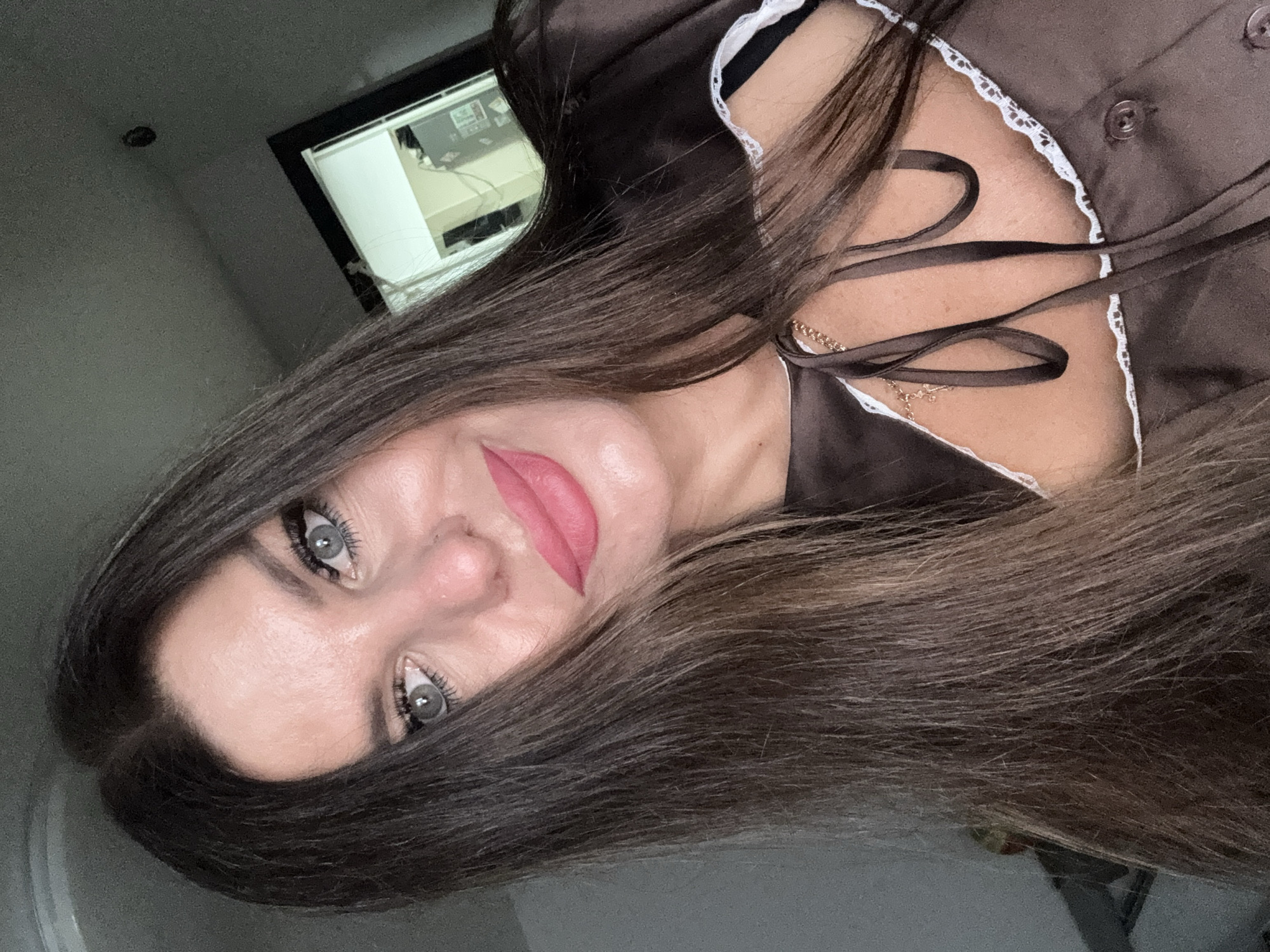 A medium close-up, head-and-shoulders shot shows a woman with long, dark brown hair and pale skin. She is looking directly at the camera with light blue eyes and her lips are painted a bright pink. She is wearing a dark brown, satin top with lace trim and dark ribbon ties at the neckline. A delicate gold chain with a small cross pendant is visible around her neck. The background is out of focus and appears to be an indoor setting, possibly a room with a window or mirror reflecting some light. There are no discernible landmarks or features that would suggest a specific location in Chaikovsky, Russia.