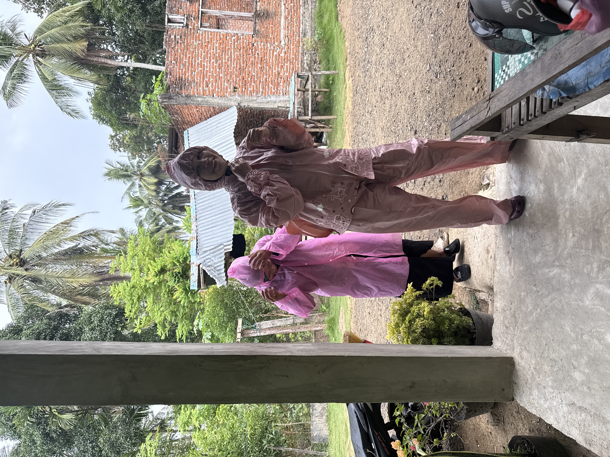Two individuals, both wearing pink raincoats and hoods, are standing on a concrete porch with a gravel yard and a brick building in the background. The person on the right, closer to the camera, is smiling and adjusting their hood. The person on the left is facing away from the camera and appears to be adjusting their own hood. Palm trees and various green foliage are visible in the background. A motorcycle is parked to the left of the porch, and potted plants are situated near the entrance. A wooden bench with a helmet on it is visible in the yard. The scene appears to be located in a rural or semi-rural area in Babahjurong, Indonesia, likely during a rainy or overcast day.